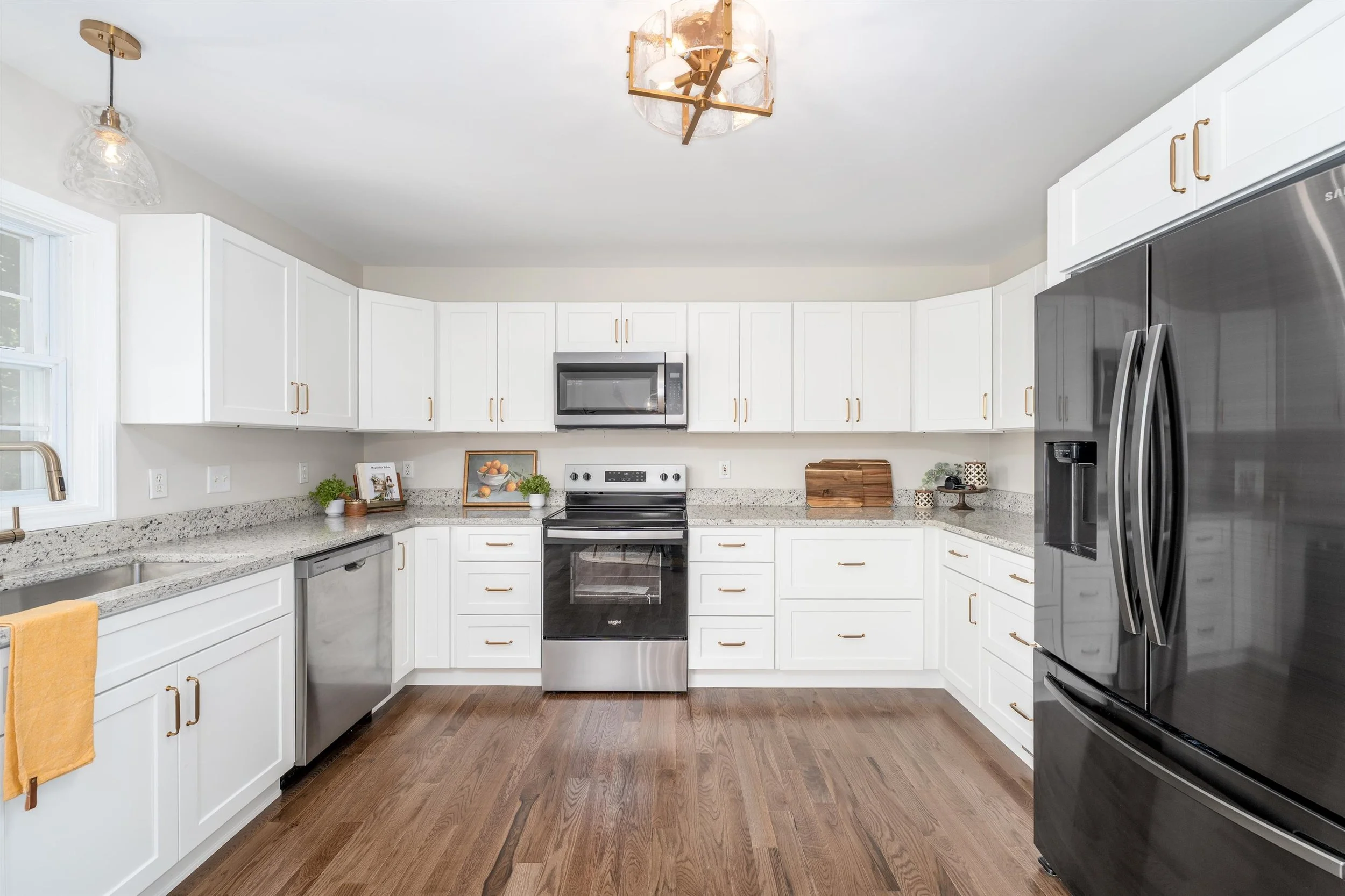 A modern kitchen with white cabinets, granite countertops, and stainless steel appliances including a microwave and refrigerator. There are decorative items and a window to the left.