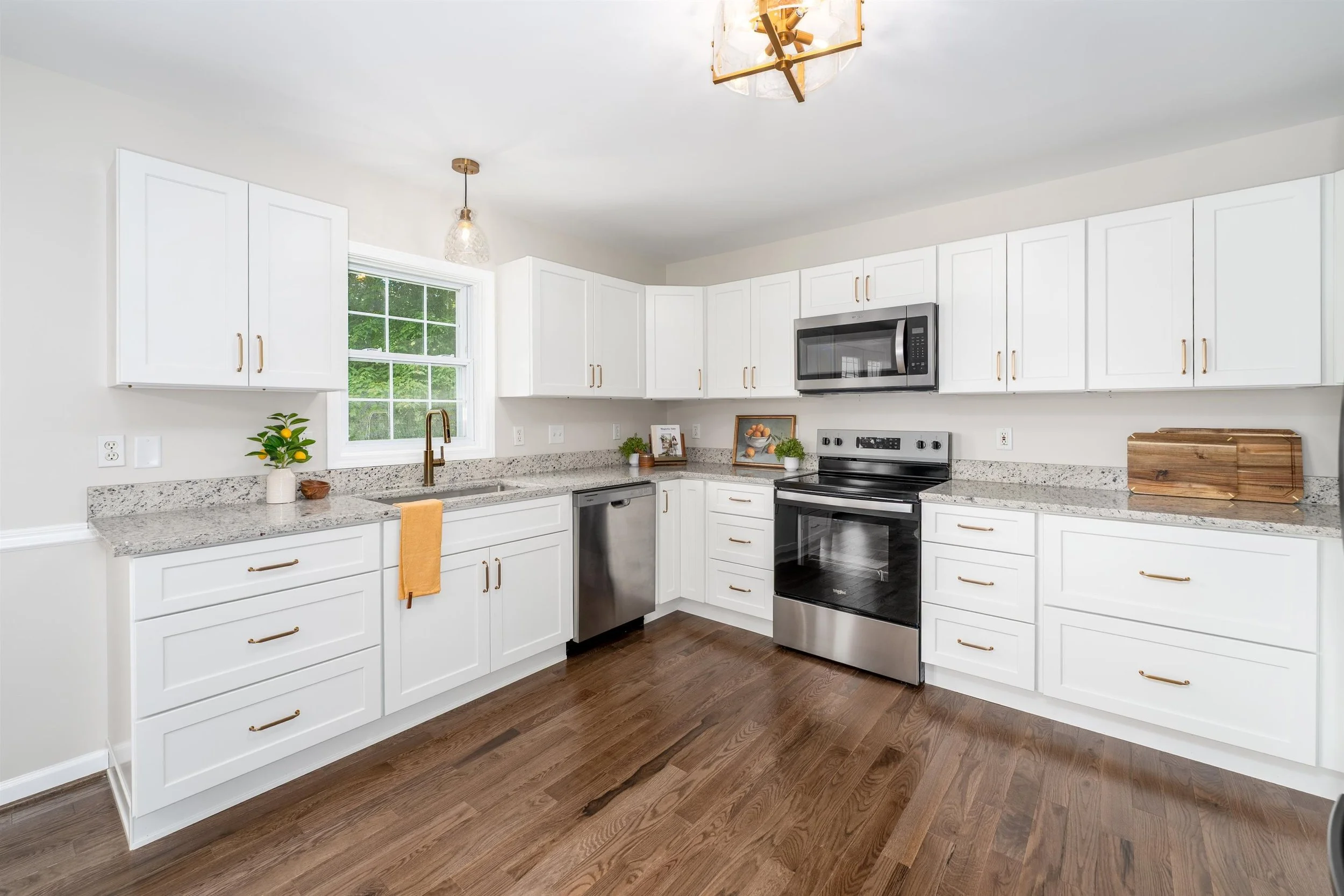 A modern kitchen with white cabinets, granite countertops, and stainless steel appliances including a microwave and dishwasher. There is a window above the sink, small potted plants, and a wooden cutting board on the counter. The flooring is dark har