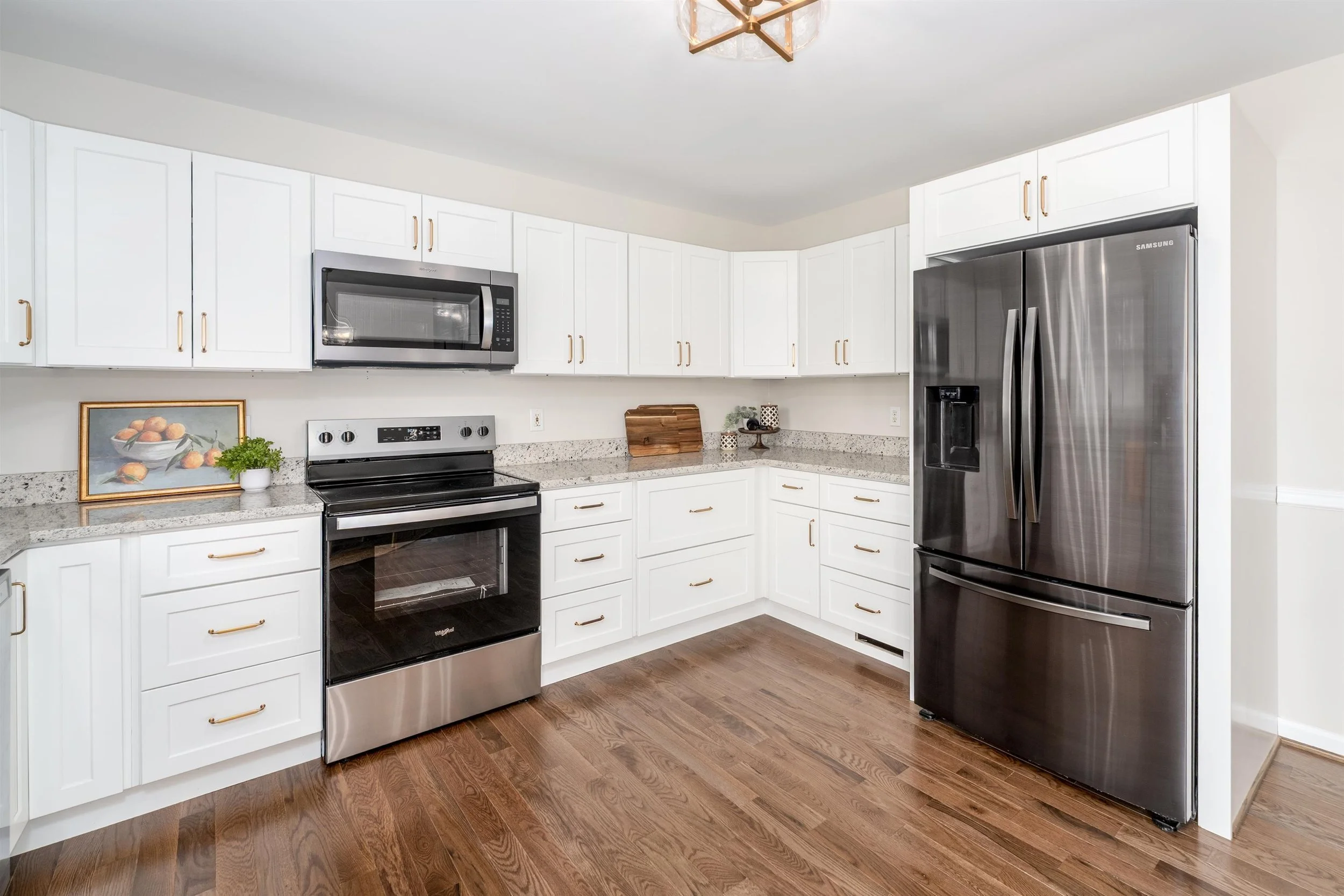 Kitchen with white cabinets, stainless steel microwave, black and stainless steel oven, and stainless steel refrigerator, with wooden floor and a framed artwork of oranges on the counter.