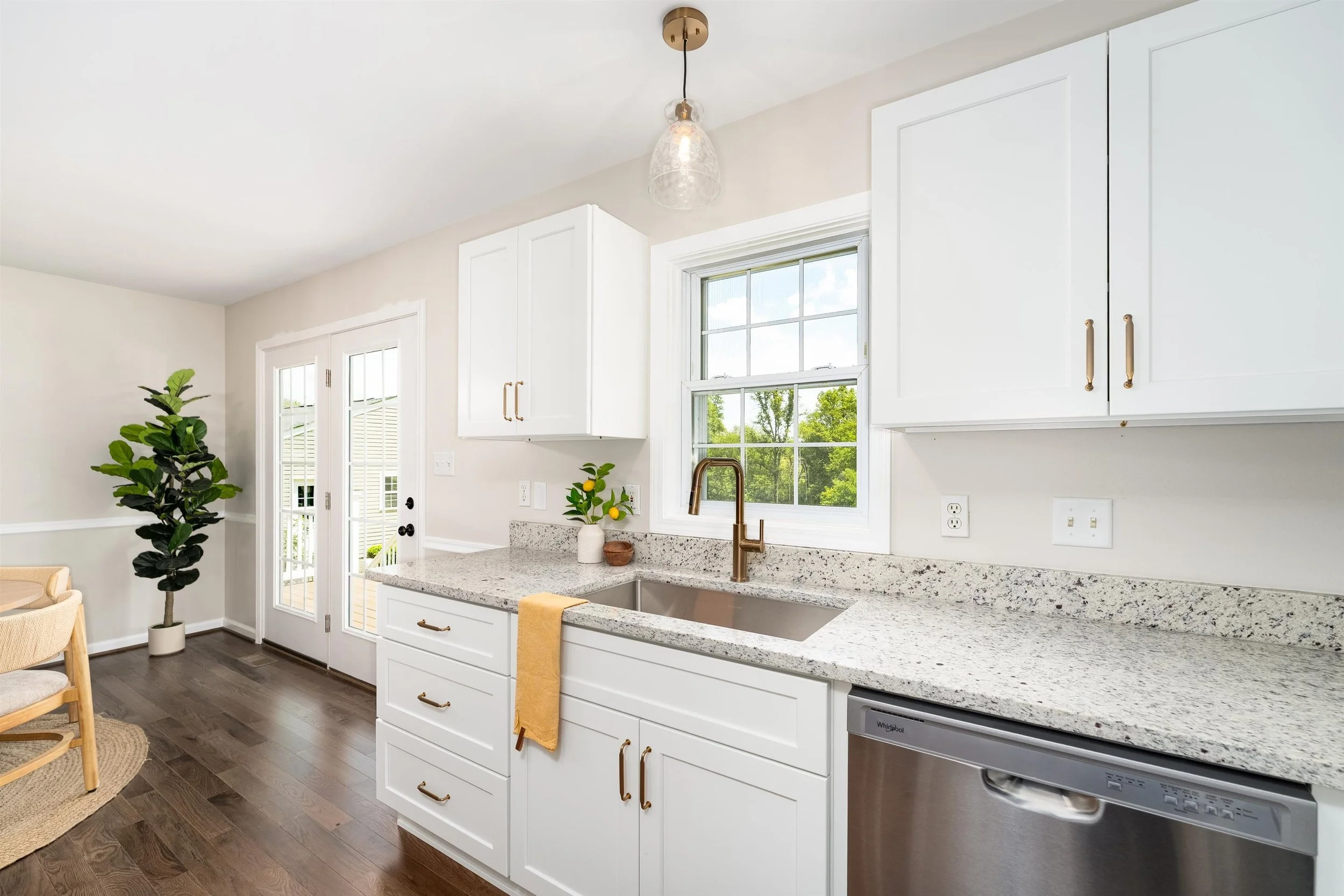Kitchen with white cabinets, granite countertop, stainless steel sink, window showing greenery outside, hanging light fixture, potted plants, and a dishwasher.
