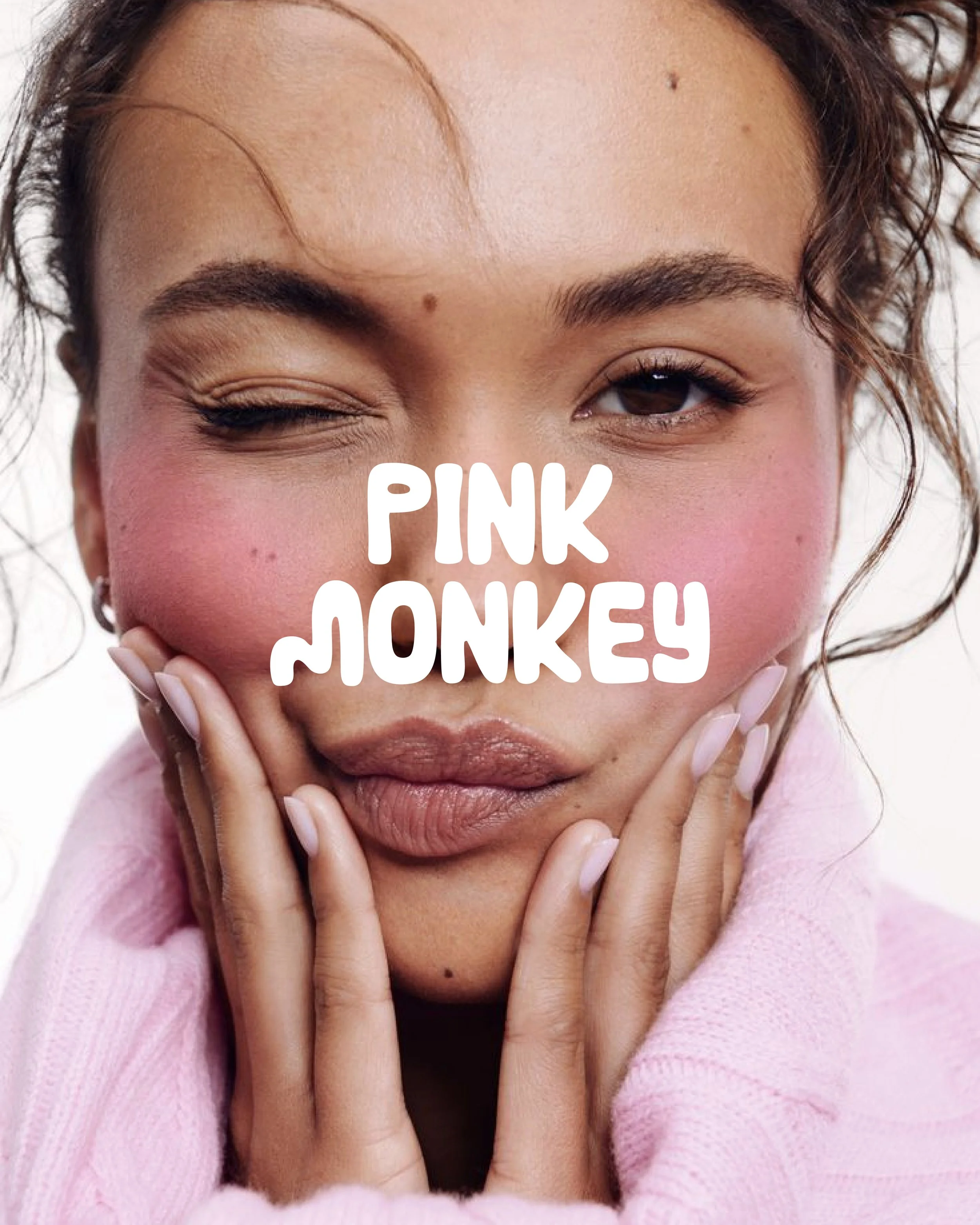 Close-up portrait of a woman with curly hair, winking with pink blush and lip color, wearing a pink sweater, with words 'Pink Monkey' over her face.
