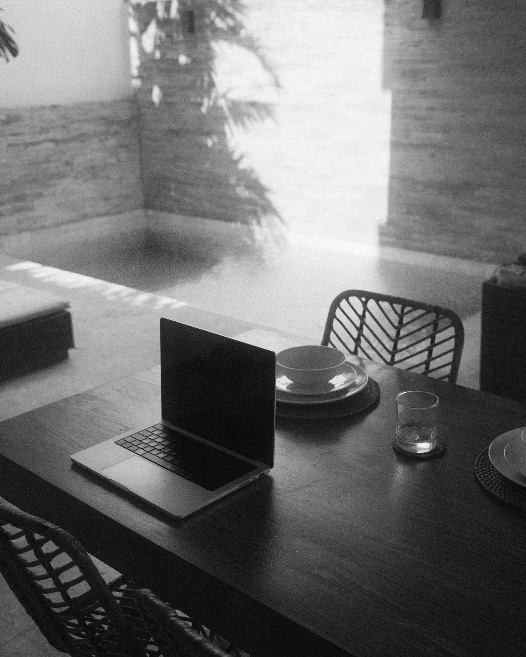 A wooden dining table with a laptop, a bowl on a plate, a glass, and a plate setup with placemats in a room with patterned chairs. Sunlight casts shadows of leaves on textured walls and appears to be coming from an outdoor pool area.