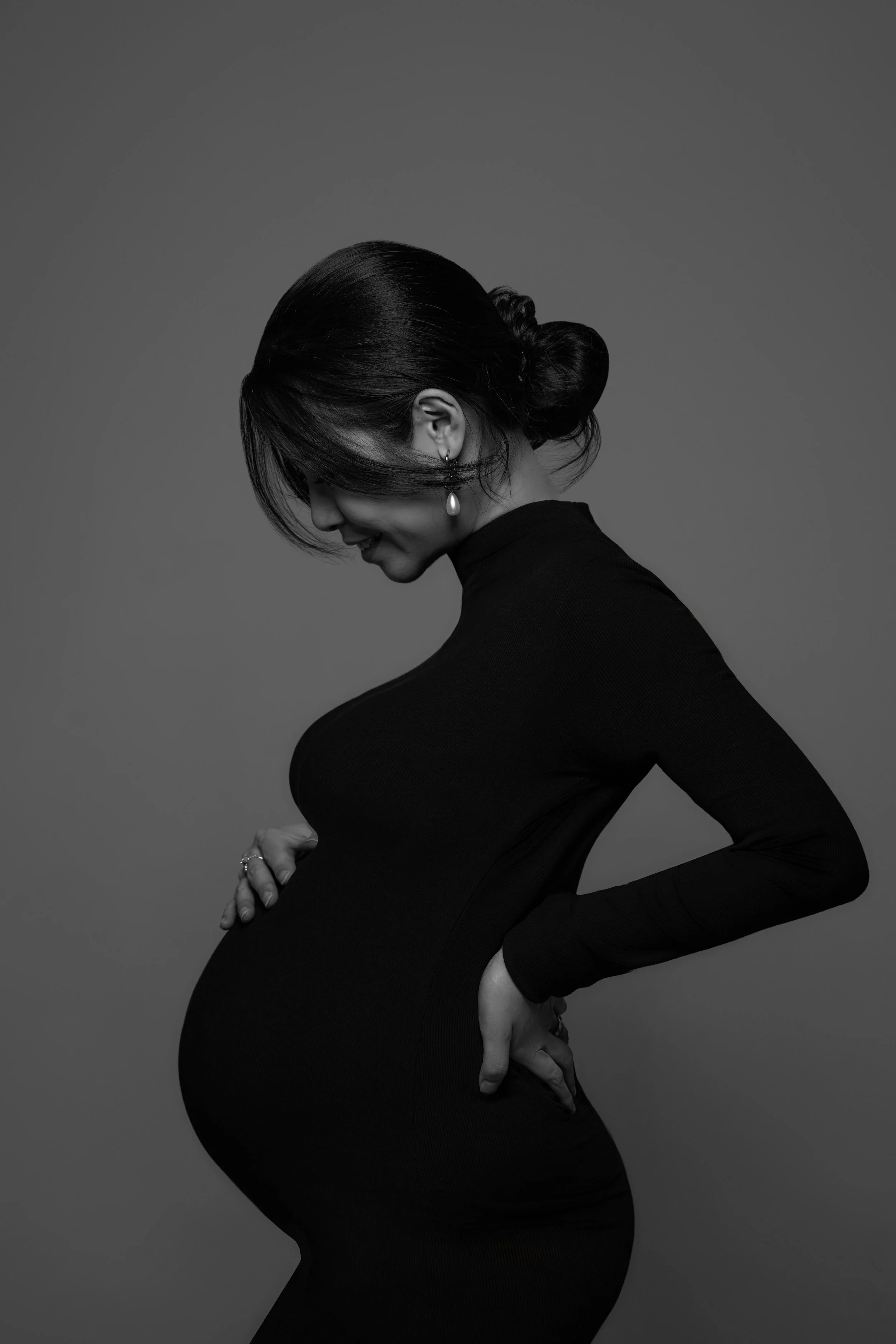 A black and white photo of a pregnant woman in profile, with her hand on her belly, wearing a long-sleeved dress and earrings, smiling softly against a plain background.