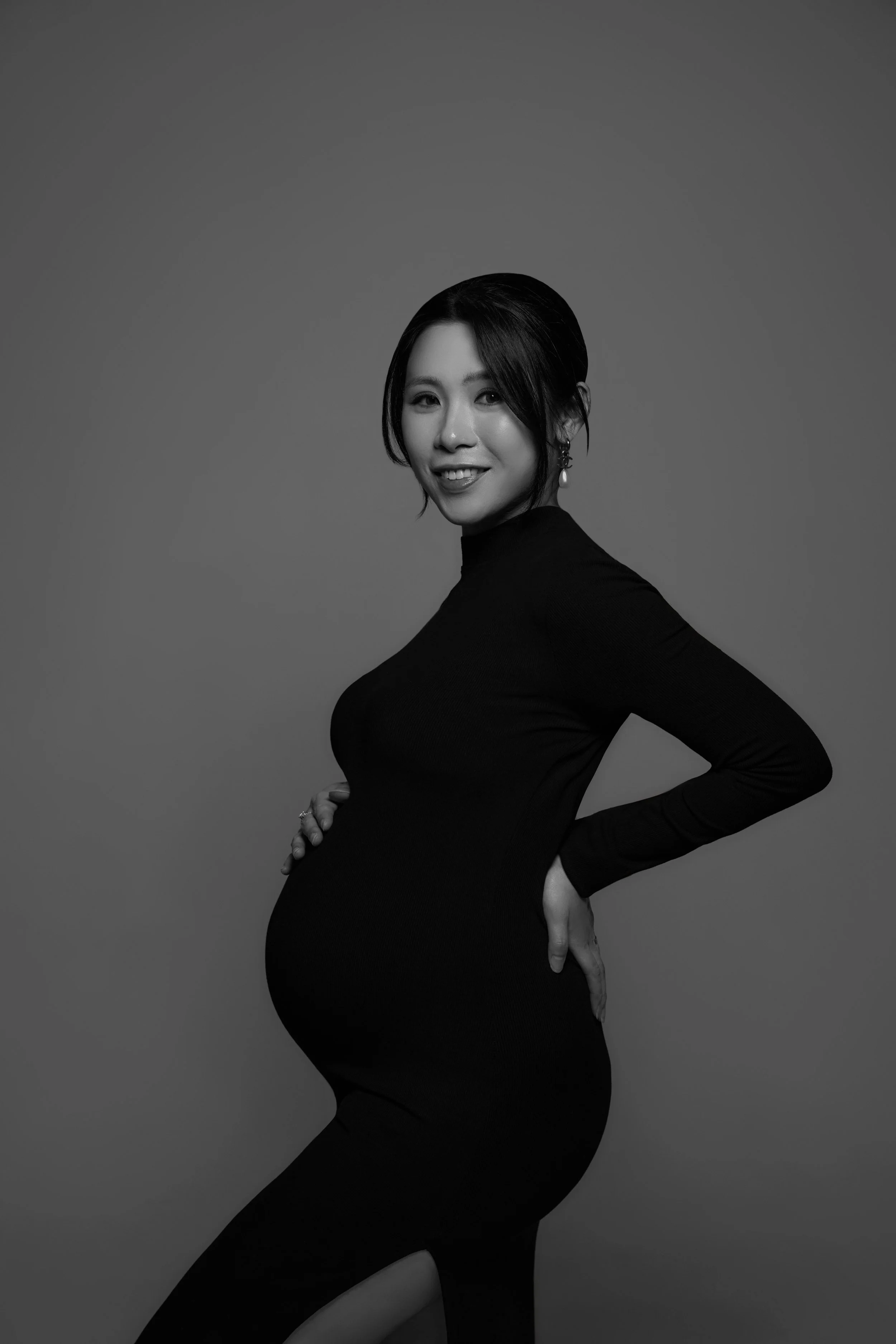 Black and white portrait of a pregnant woman with short hair, smiling, wearing a long-sleeve black dress, with one hand on her hip and the other resting on her belly.