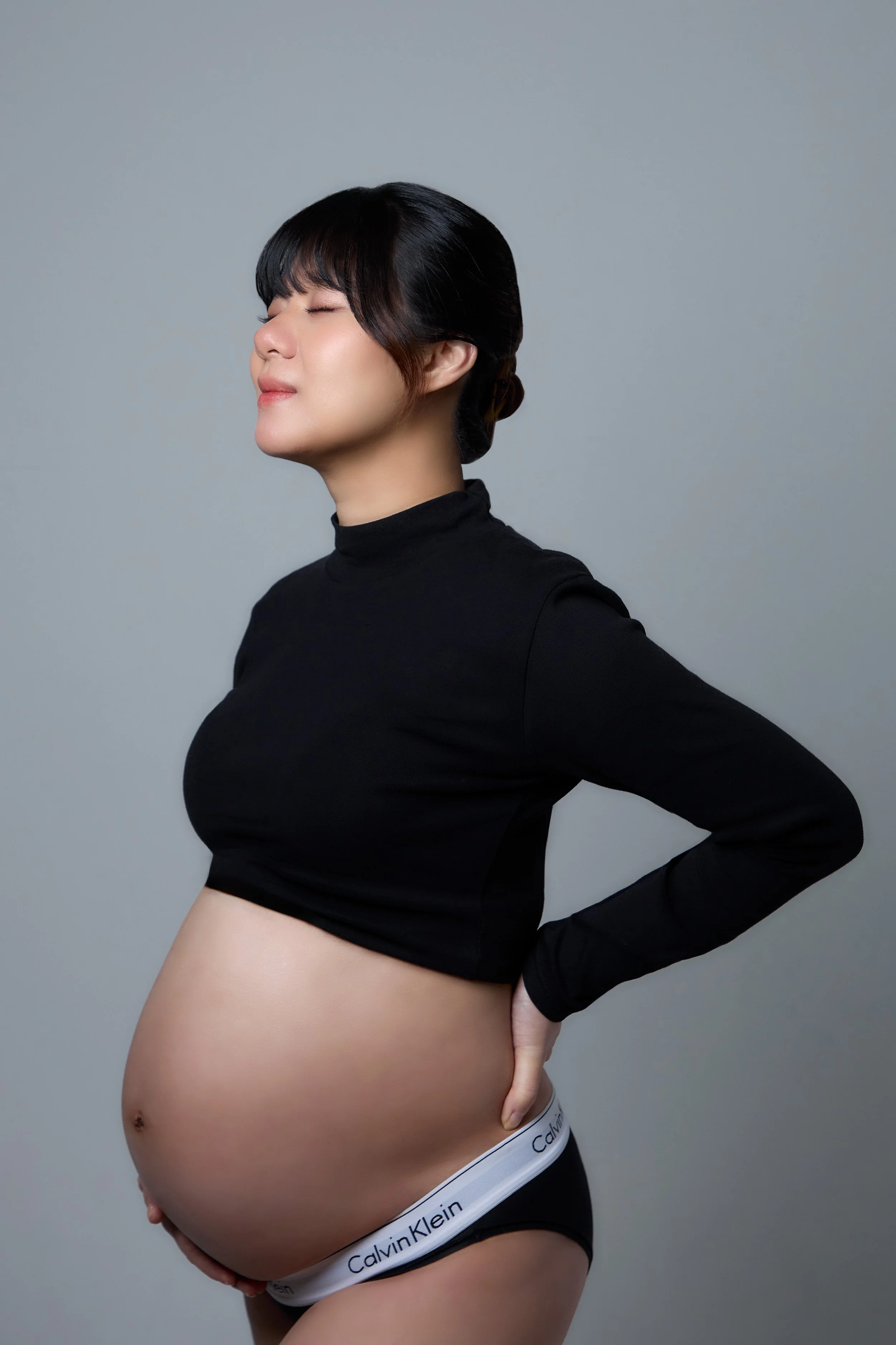 Pregnant woman with closed eyes wearing a black top and Calvin Klein underwear, standing against a gray background.