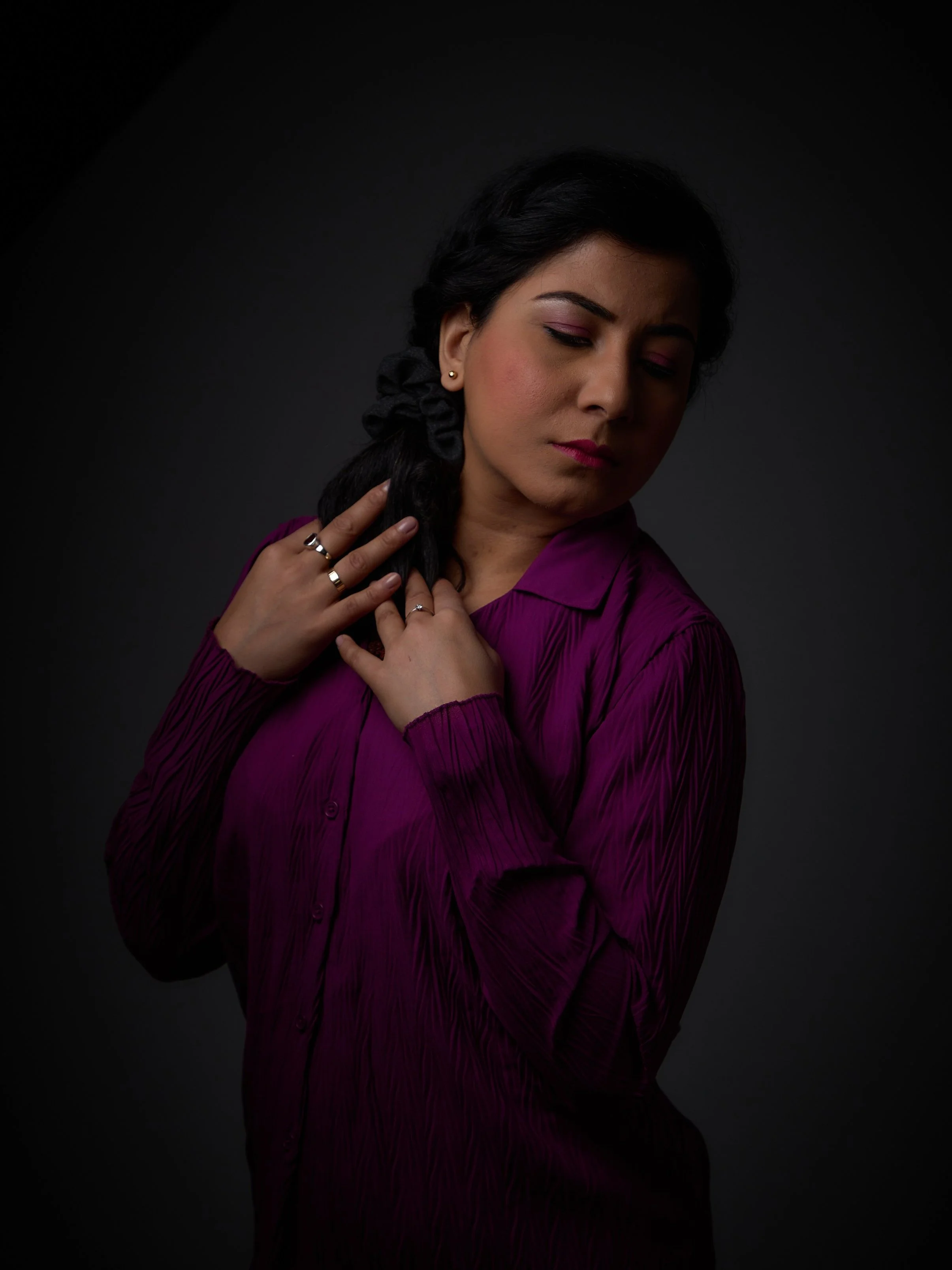 A woman with dark hair in a braid, wearing a purple blouse, is posing with her eyes closed against a dark background.