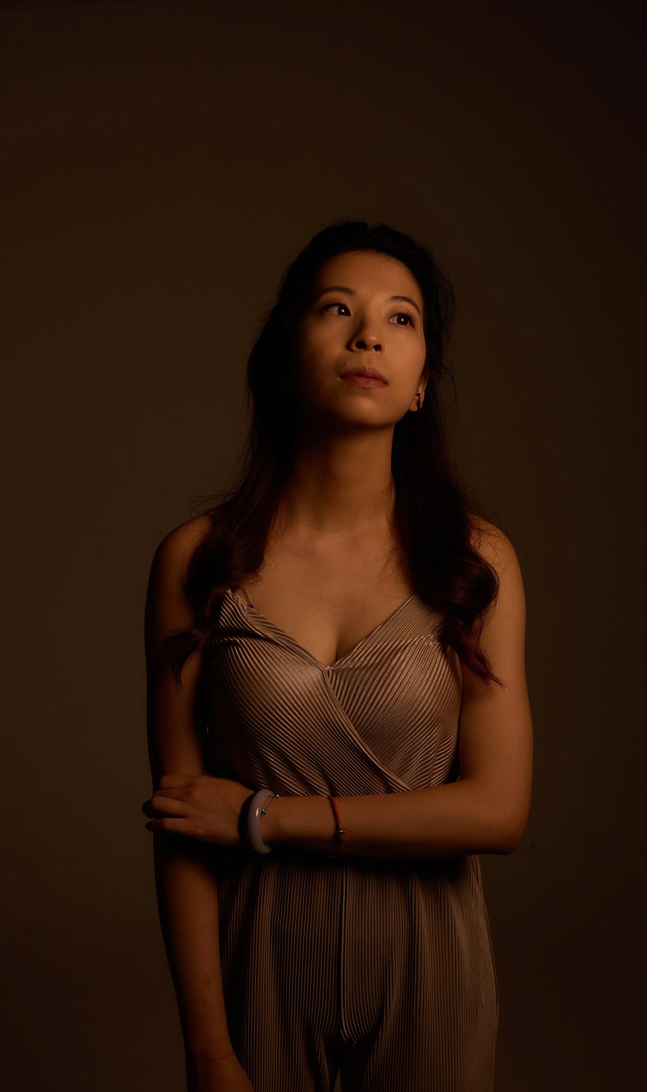 portrait of a young woman with long dark hair looking thoughtfully to the side, wearing a striped sleeveless top and bracelets on her wrist, against a dark plain background.