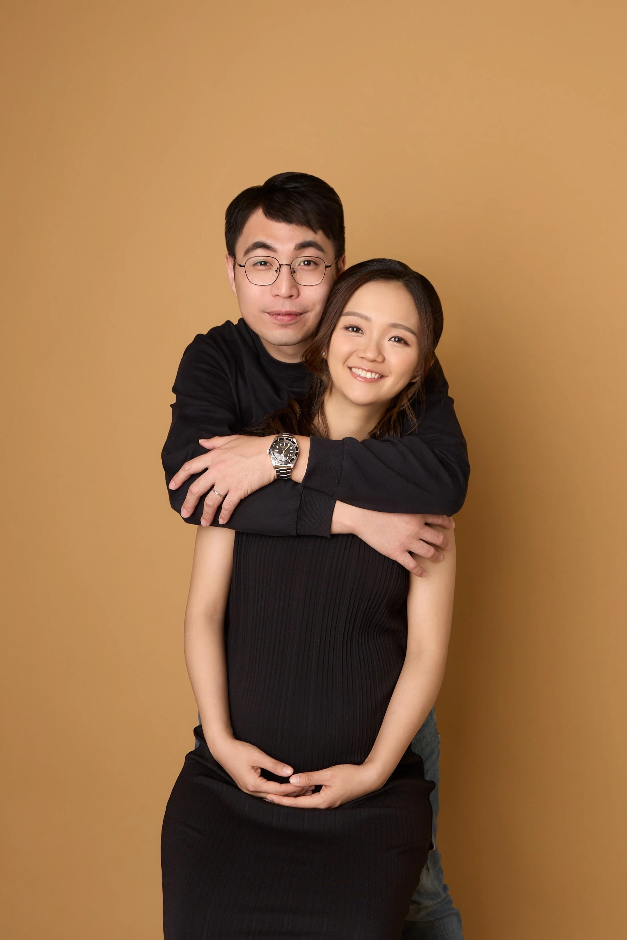 A smiling pregnant woman in a black dress is being hugged from behind by a man wearing glasses and a black long-sleeve shirt, against a plain beige background.