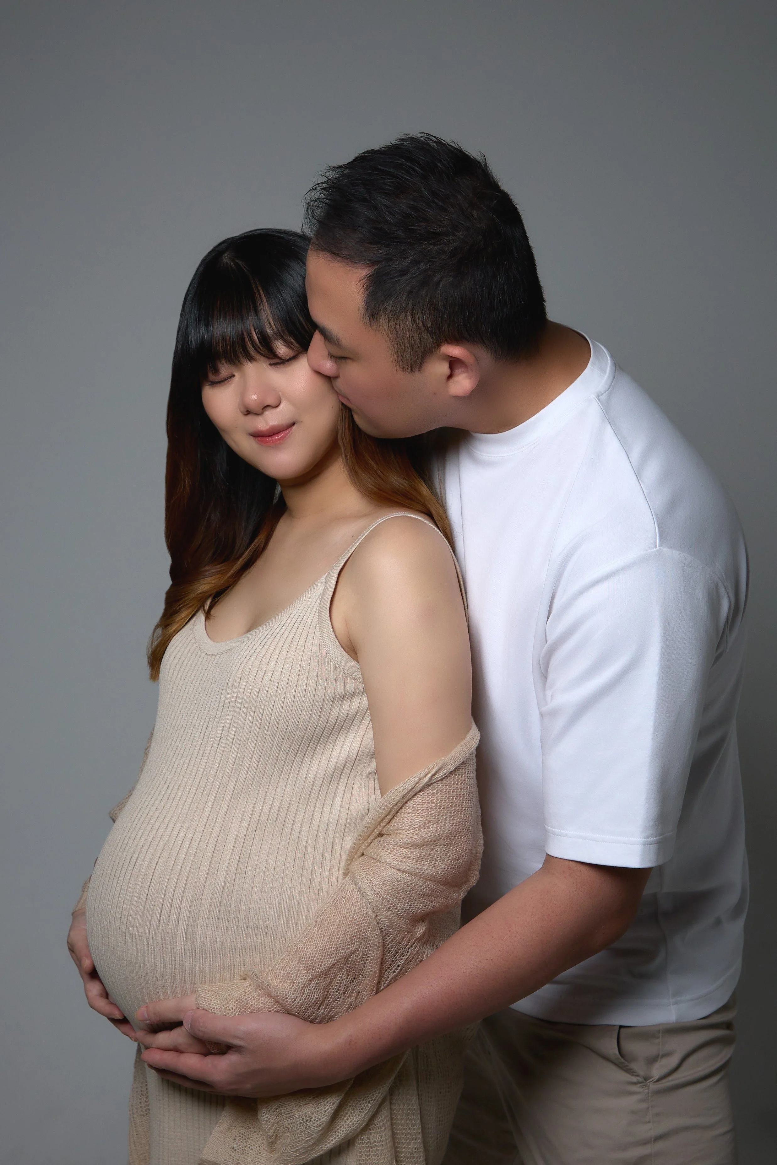 A pregnant woman with closed eyes and a serene expression is holding her belly, while a man kisses her on the forehead, both standing against a plain gray background.