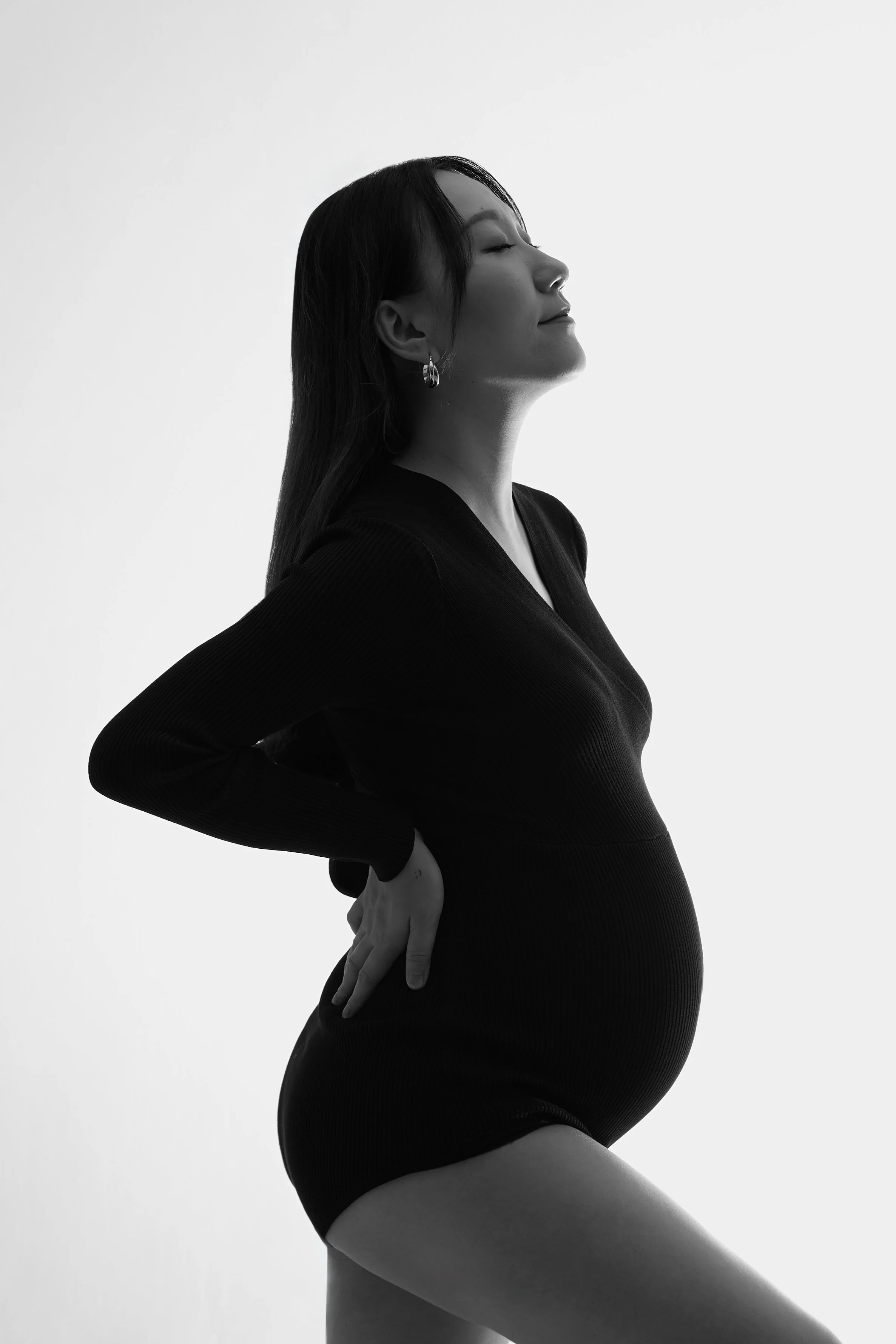 A pregnant woman in a black long-sleeve top and shorts, standing sideways with one hand on her back and her eyes closed in front of a plain wall.