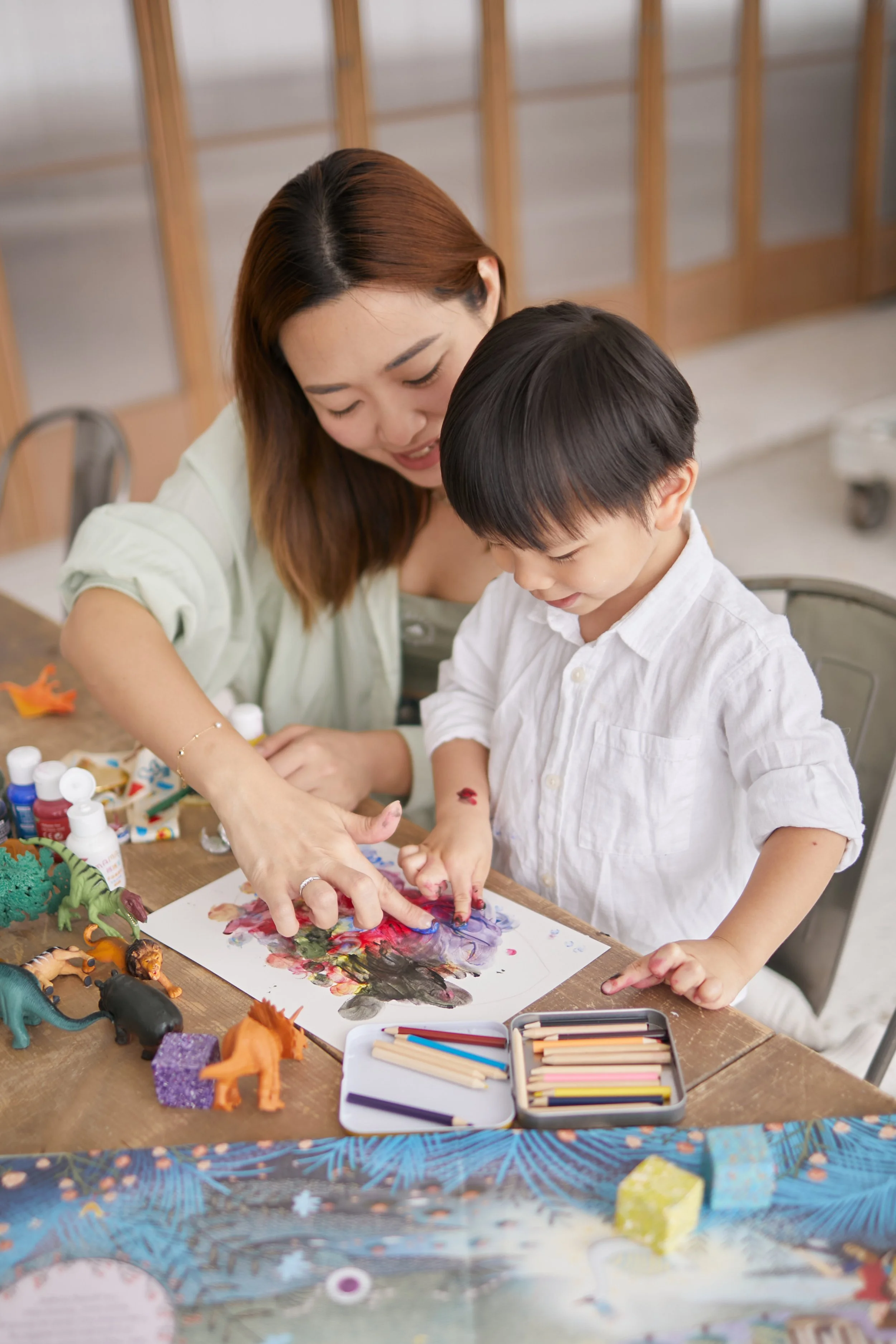 A woman and a young boy engaging in a creative art project at a table, using paints, colored pencils, and toy dinosaurs.