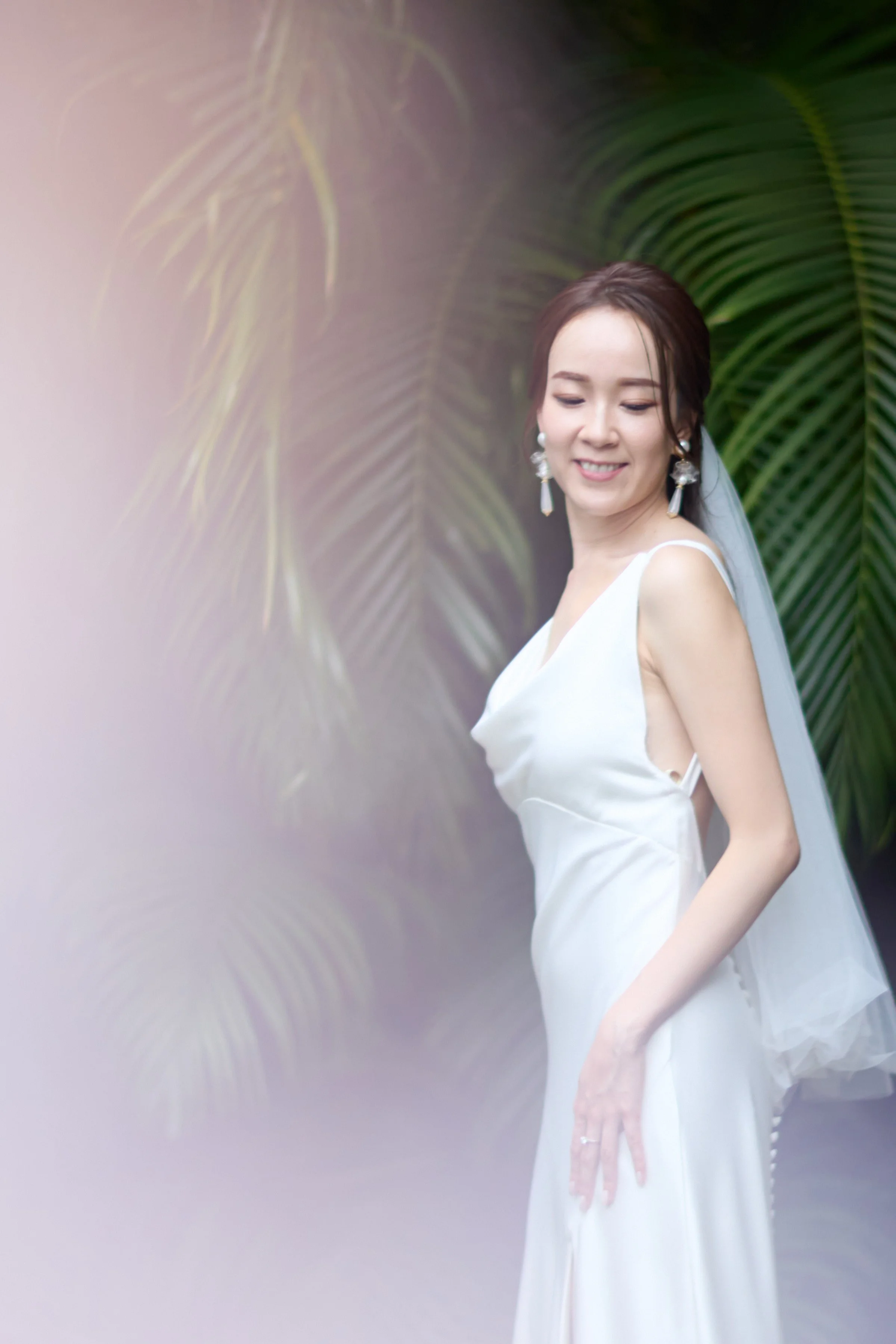 A bride in a white wedding gown with a veil, standing outdoors near green palm leaves, smiling softly.