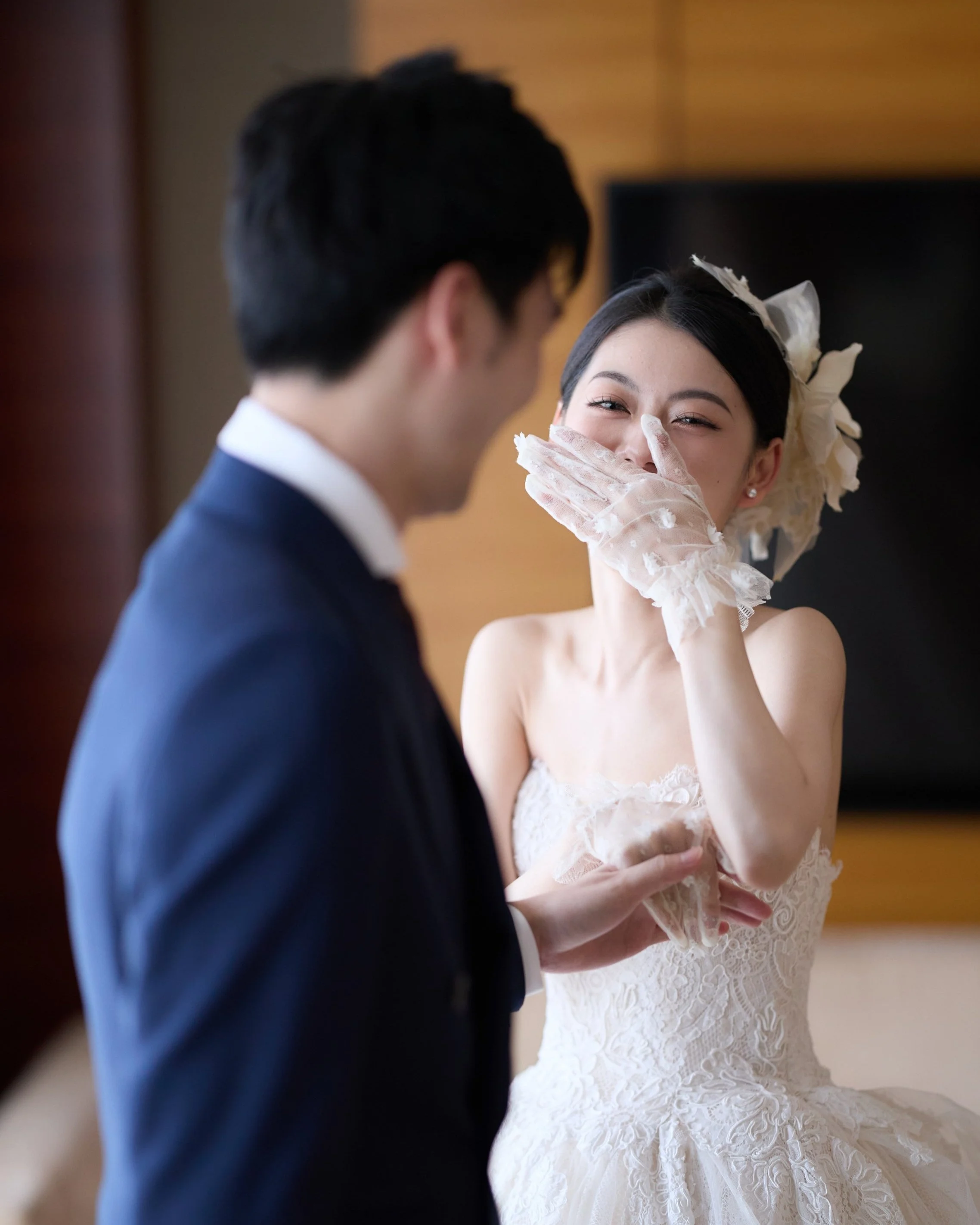 A bride in a wedding dress laughing and covering her mouth with her gloved hand, while a groom in a suit faces her in a warmly lit room.