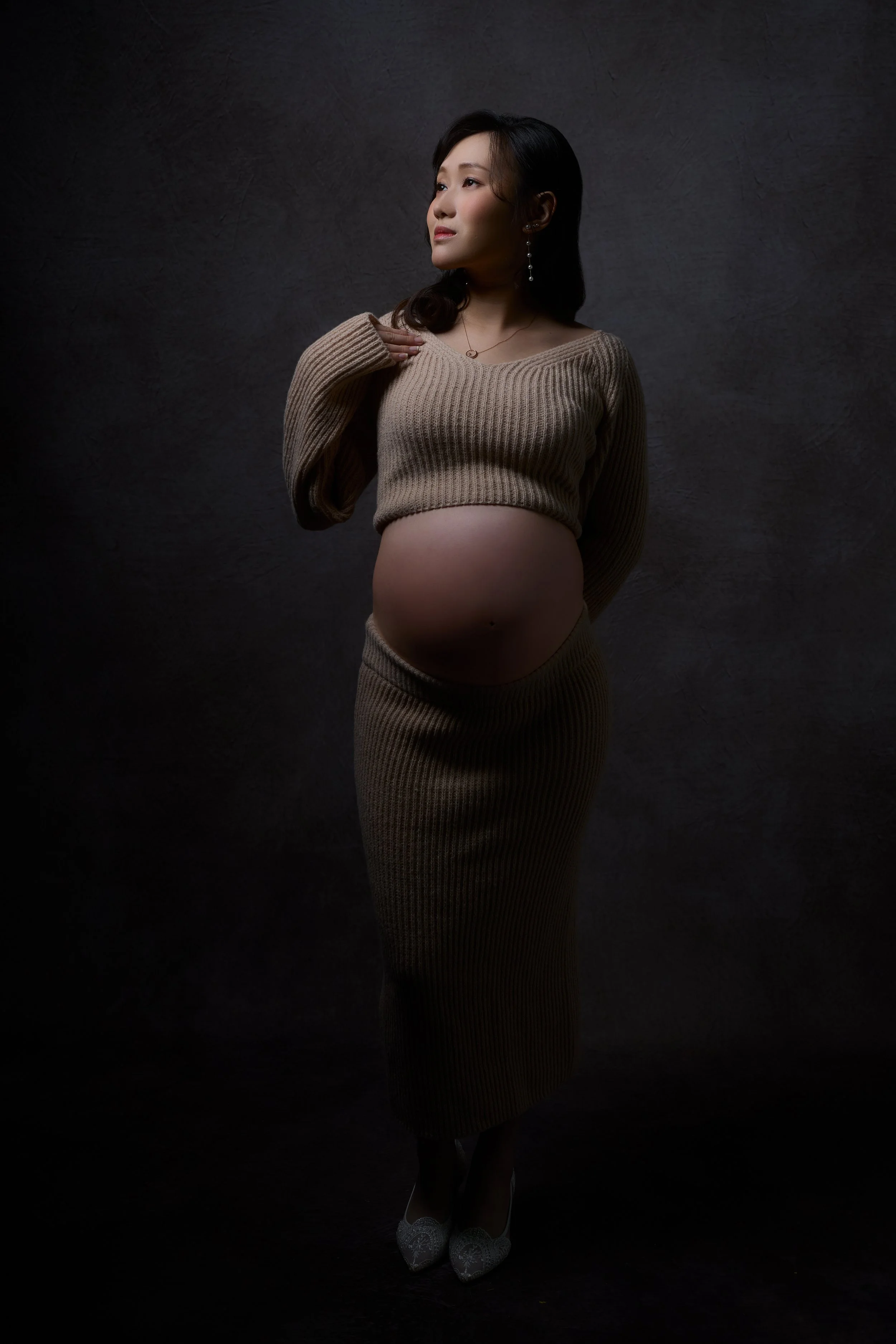 A pregnant woman wearing a beige knitted crop top and matching skirt, standing against a dark textured background, with jewelry and silver shoes.