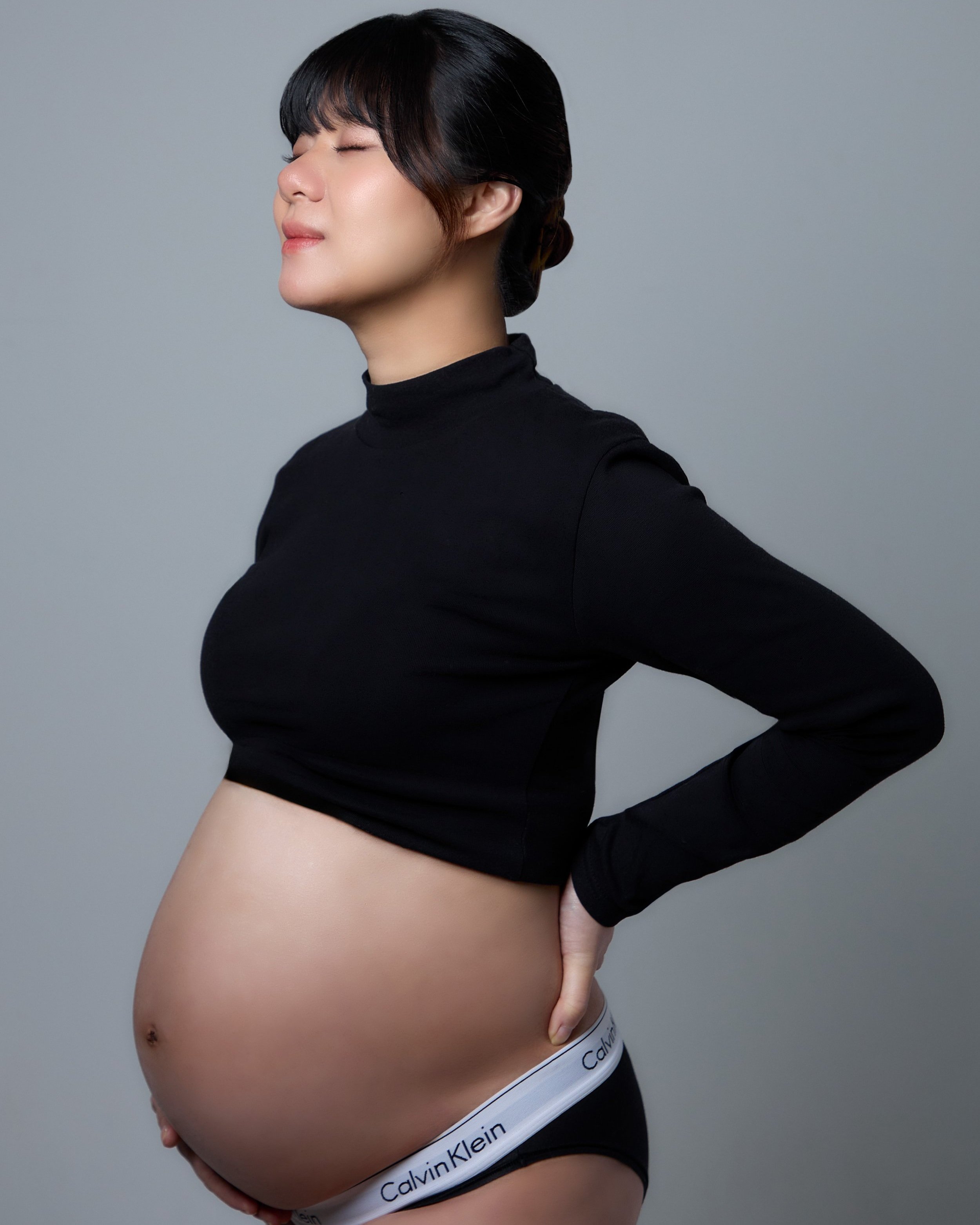 Pregnant woman with black hair in a bun, wearing a black turtleneck and Calvin Klein underwear, standing with her eyes closed and hand on her lower back, against a gray background.