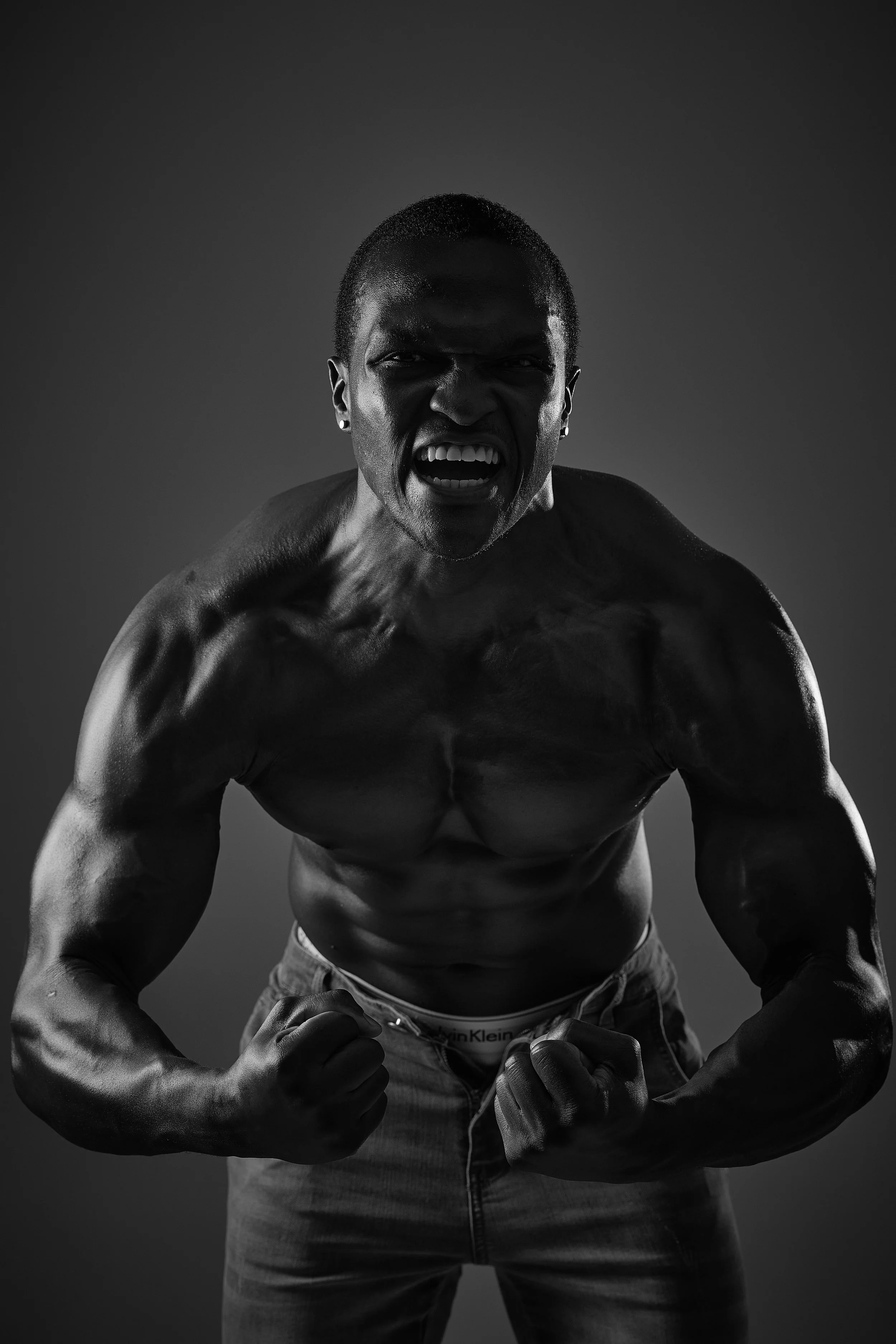 A muscular man fist-pumping and shouting passionately, shirtless, with a dark background, black and white photograph.