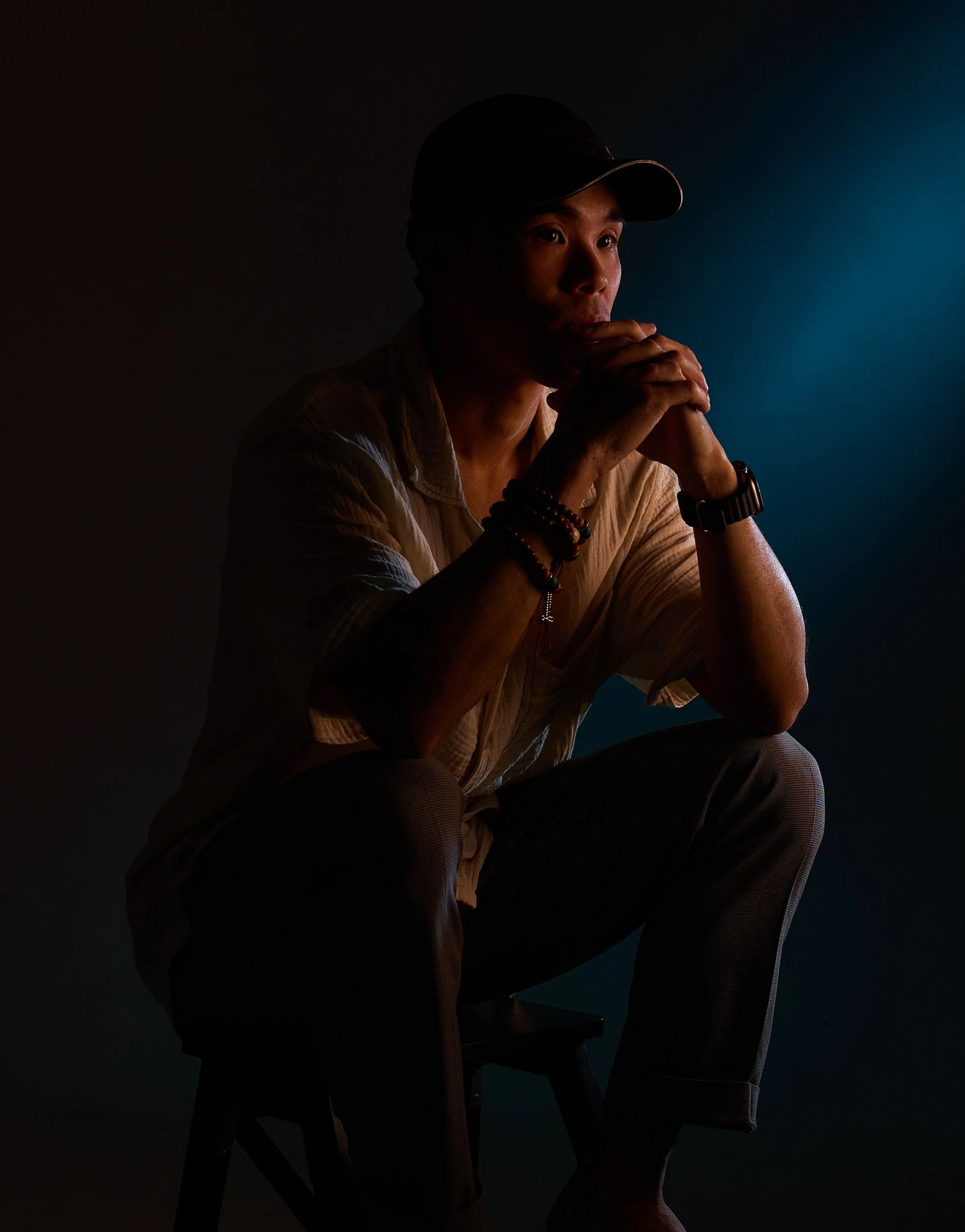 A man sitting on a stool, dressed casually, wearing a cap and multiple bracelets, appears to be deep in thought with his hands clasped near his mouth, illuminated by a soft light against a dark background.