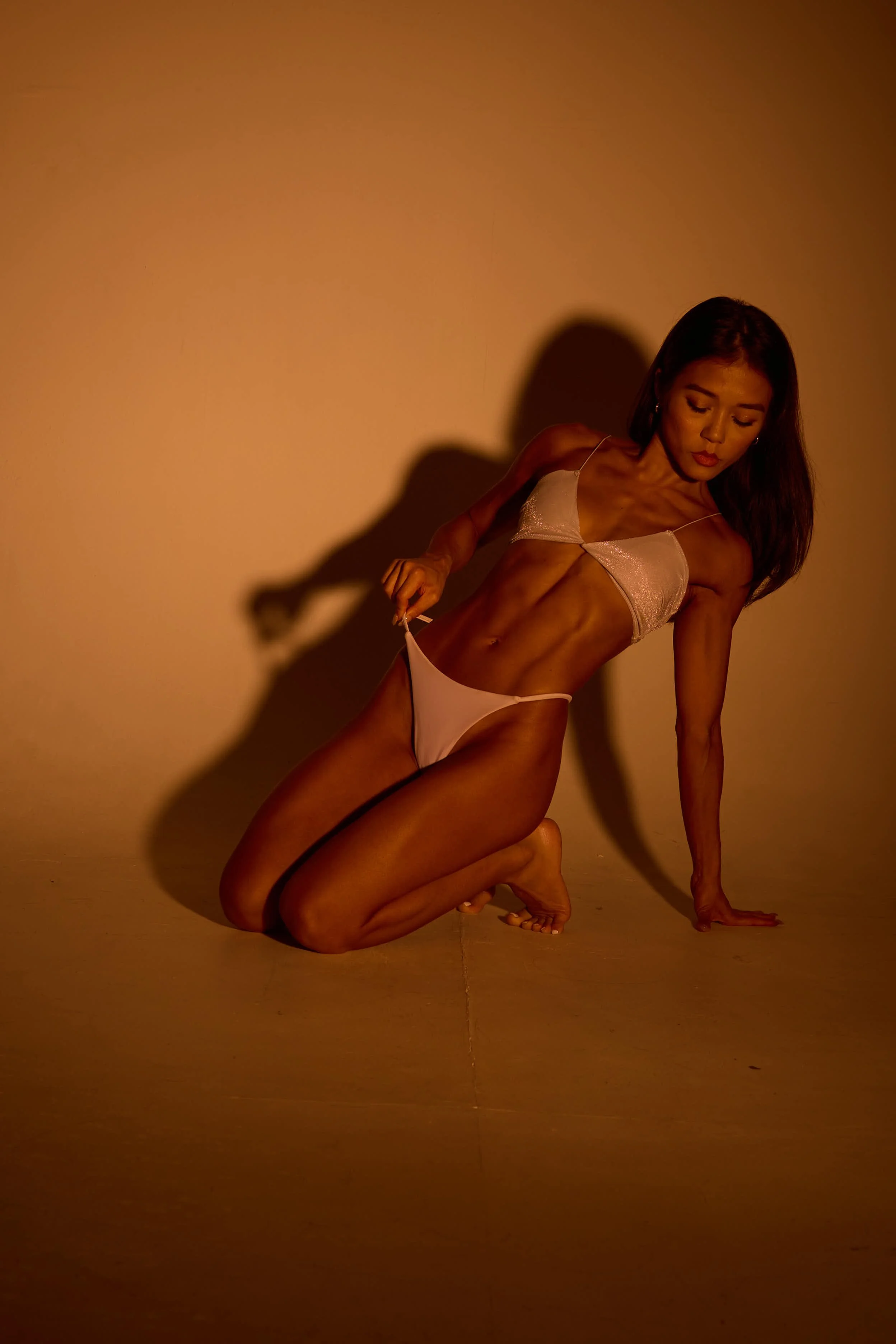 A woman with toned muscles kneeling on the floor in a dimly lit room, wearing white lingerie, casting a shadow on the wall behind her.
