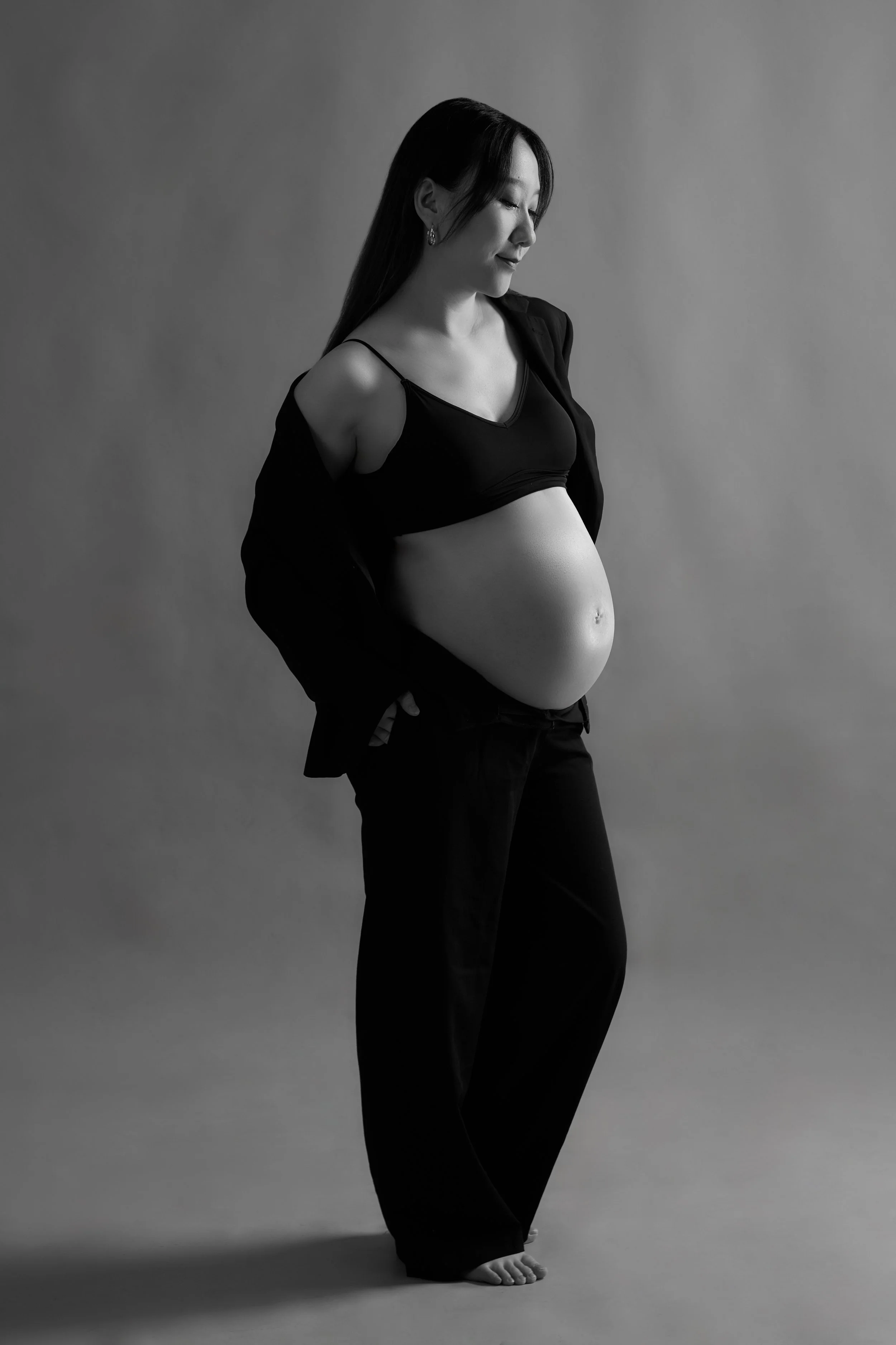 Black and white photo of a pregnant woman standing with her eyes closed, hands in her pockets, wearing a black sports bra, loose black pants, and a black blazer draped over her shoulders, against a plain background.