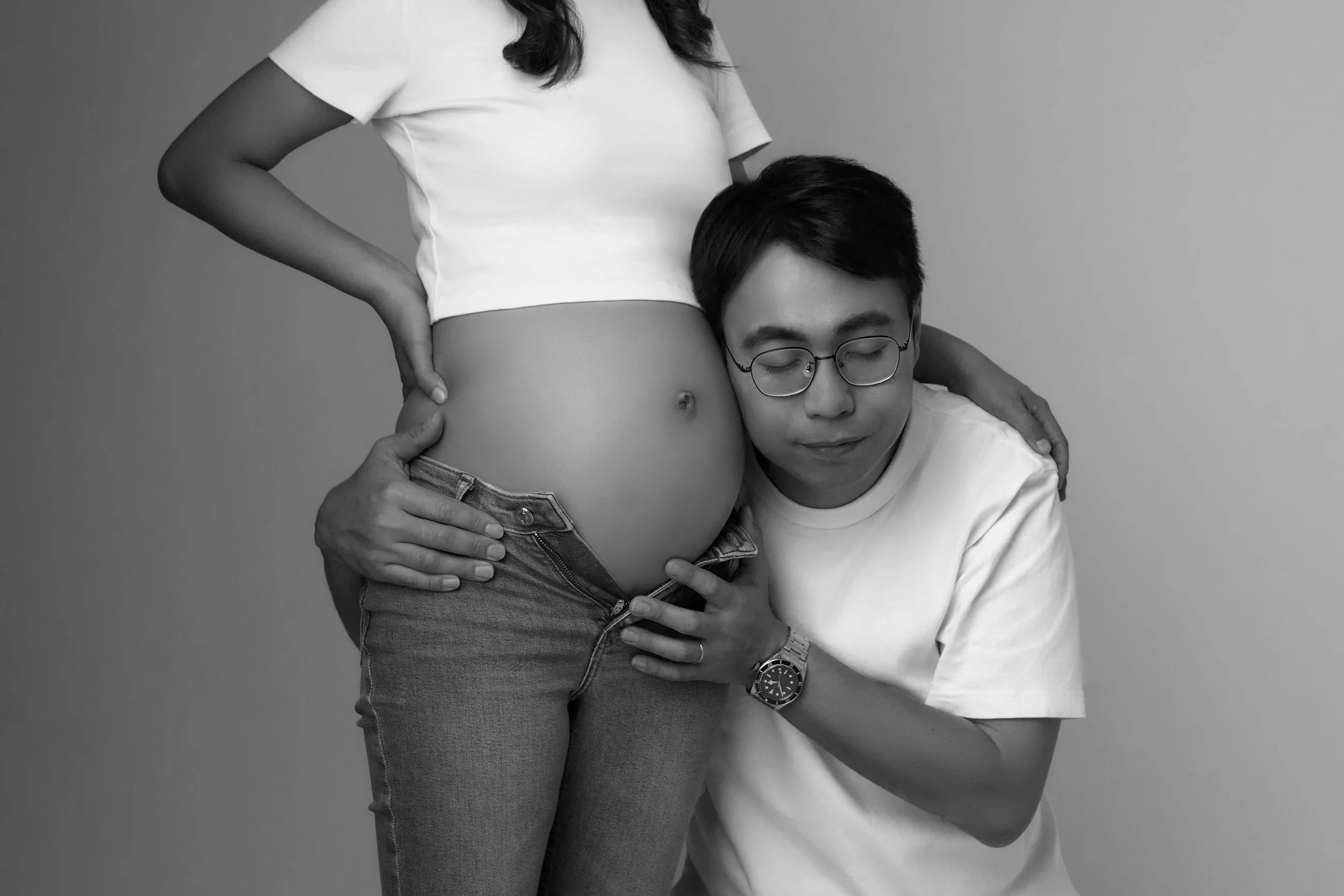 A person with a pregnant belly gently holding her belly, while another person with glasses is resting their head on her belly with eyes closed, both in a black and white photograph.