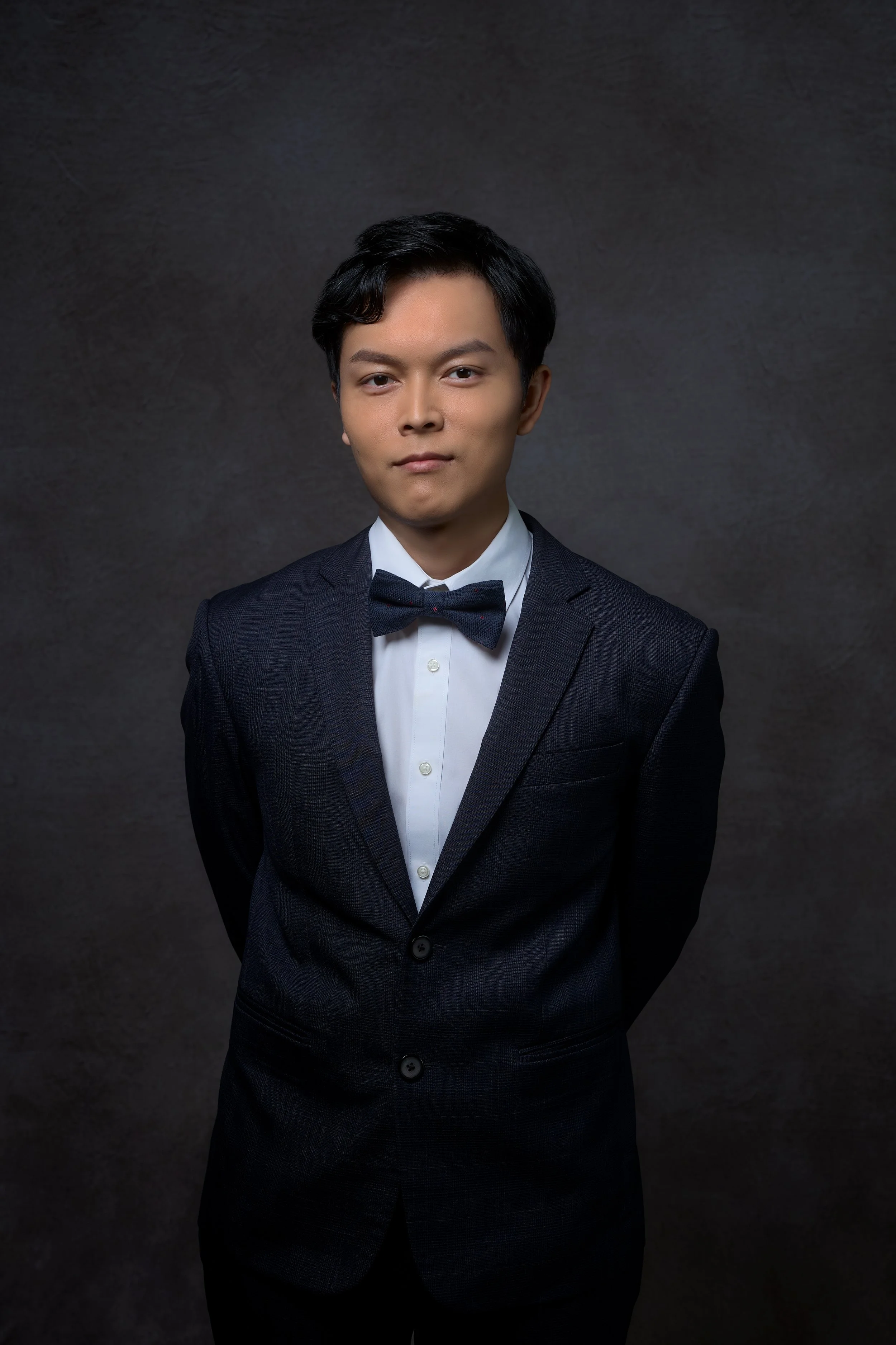 Young man in a dark suit and bow tie standing against a dark background.