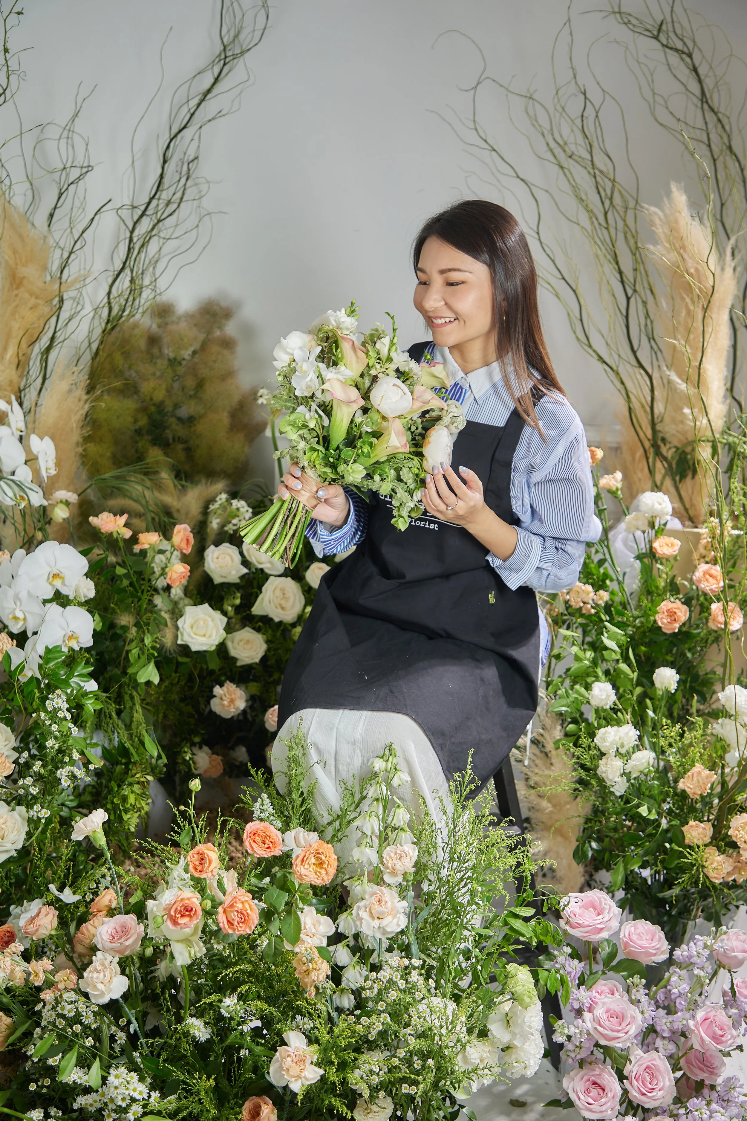 A woman in a floral shop surrounded by a variety of colorful flowers, holding a bouquet of white and pink flowers, smiling.