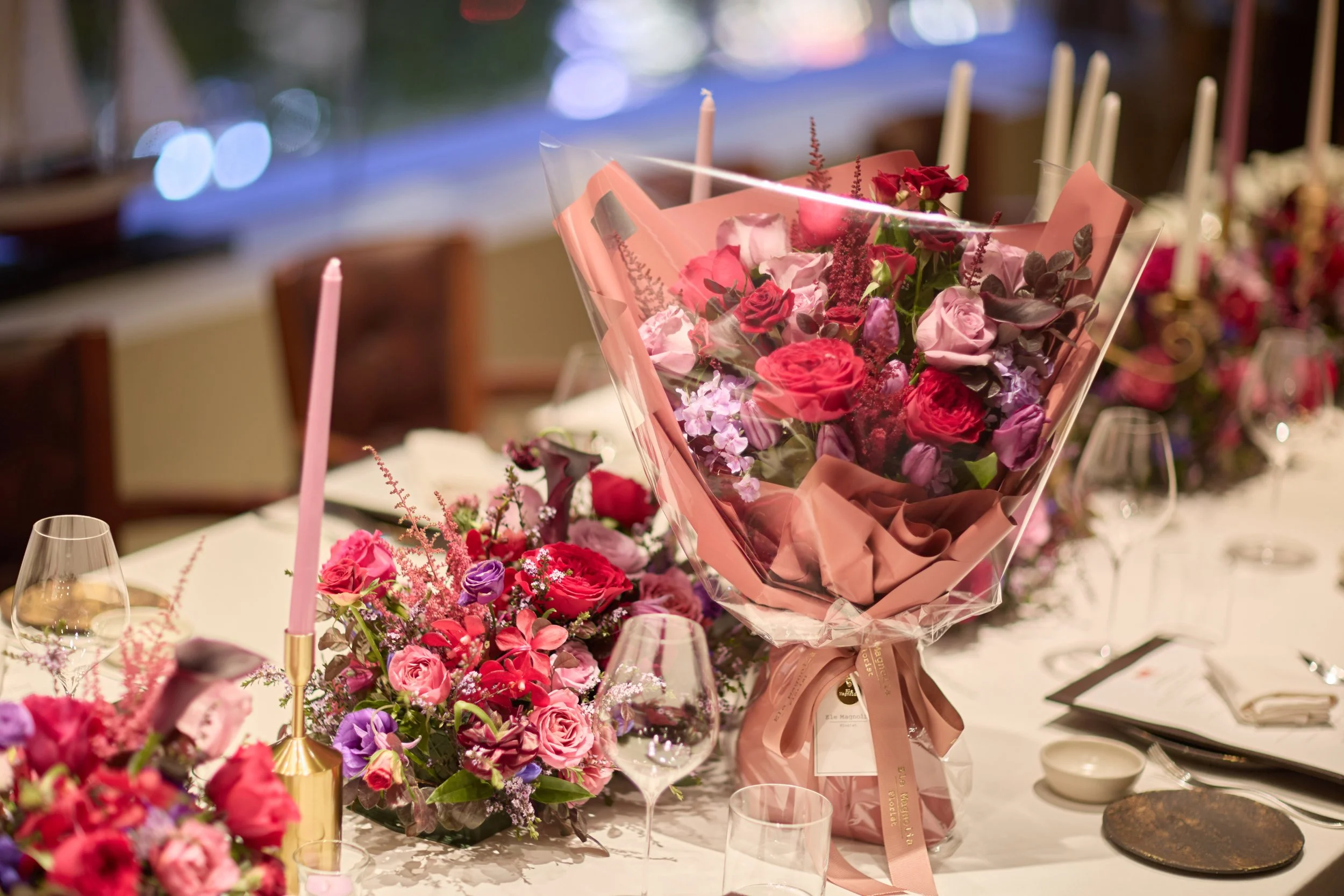 Elegant floral centerpiece with pink, red, purple, and white flowers, wrapped pink bouquet, set on a formal dining table with pink candles, wine glasses, and tableware.