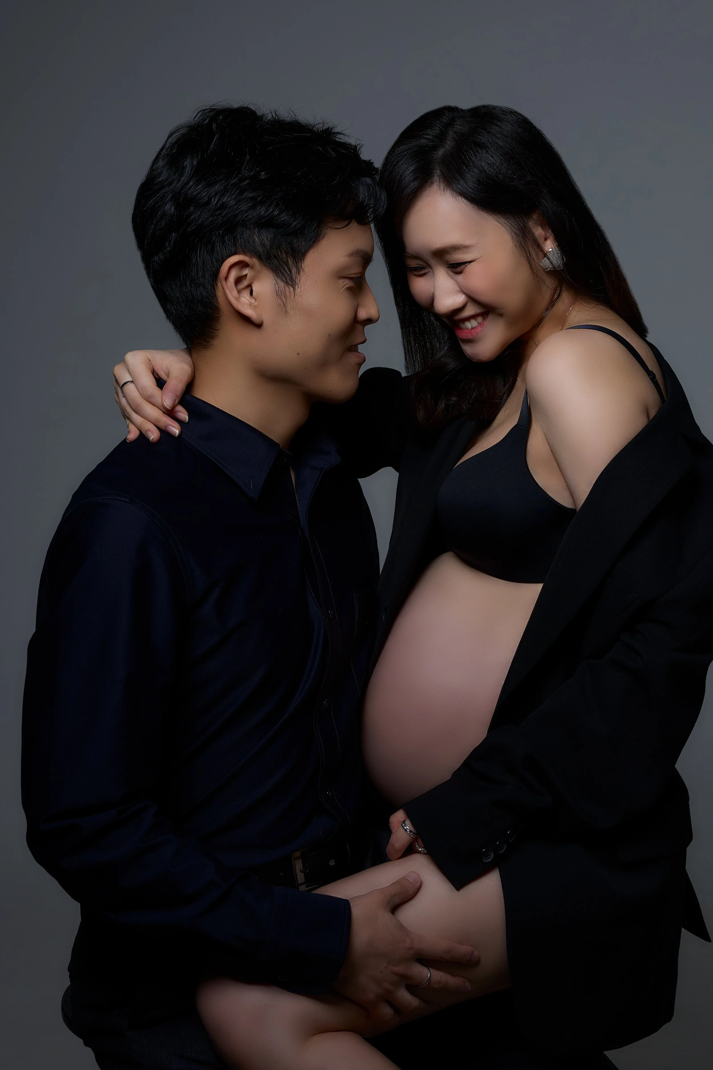 A pregnant woman and a man are smiling and touching each other affectionately in a studio setting. The woman is wearing a black bra and a black jacket off her shoulders, showing her pregnant belly. The man is wearing a dark shirt and is holding her b