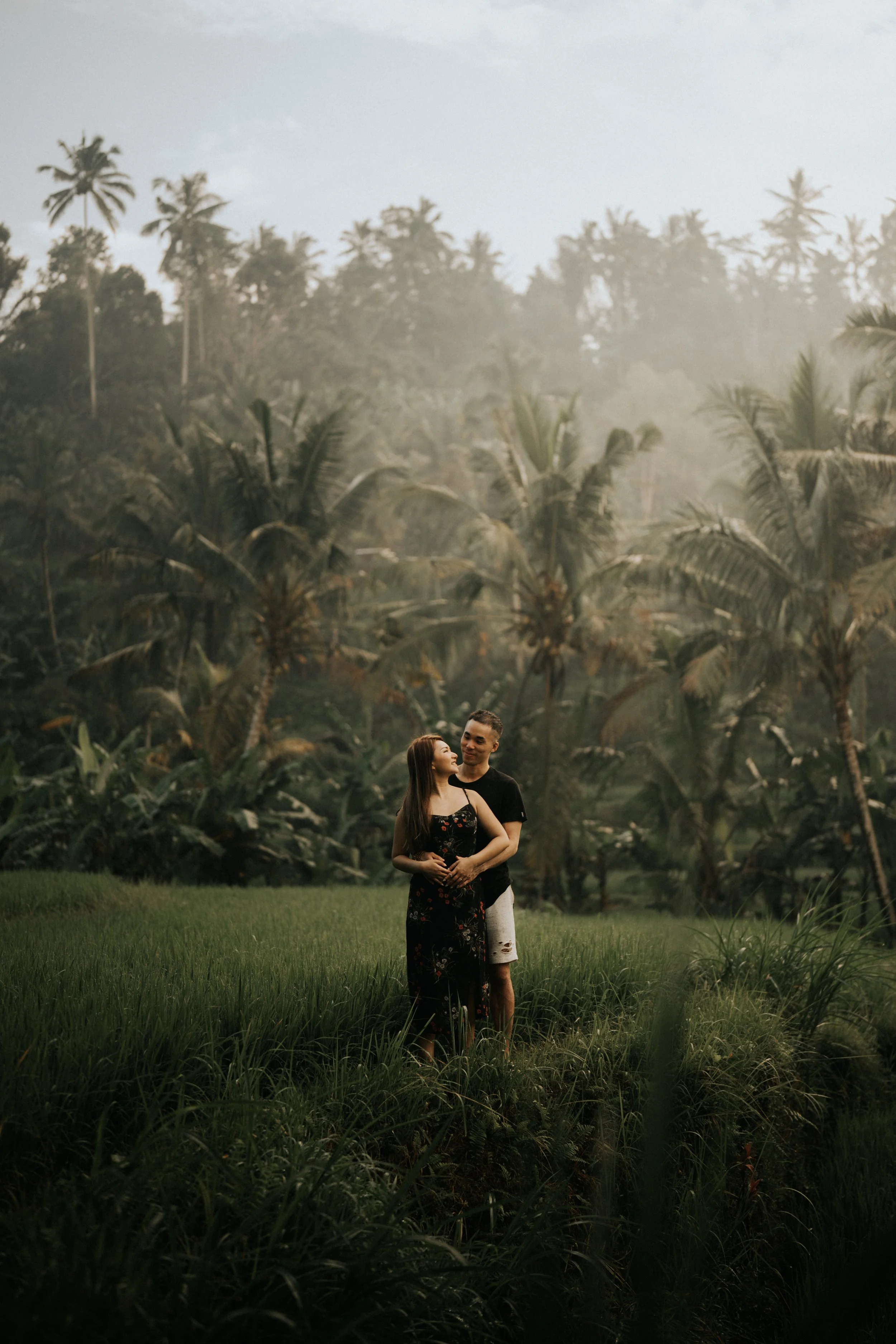 A couple standing in a lush, green field with tall palm trees and dense jungle in the background, sharing an intimate moment.