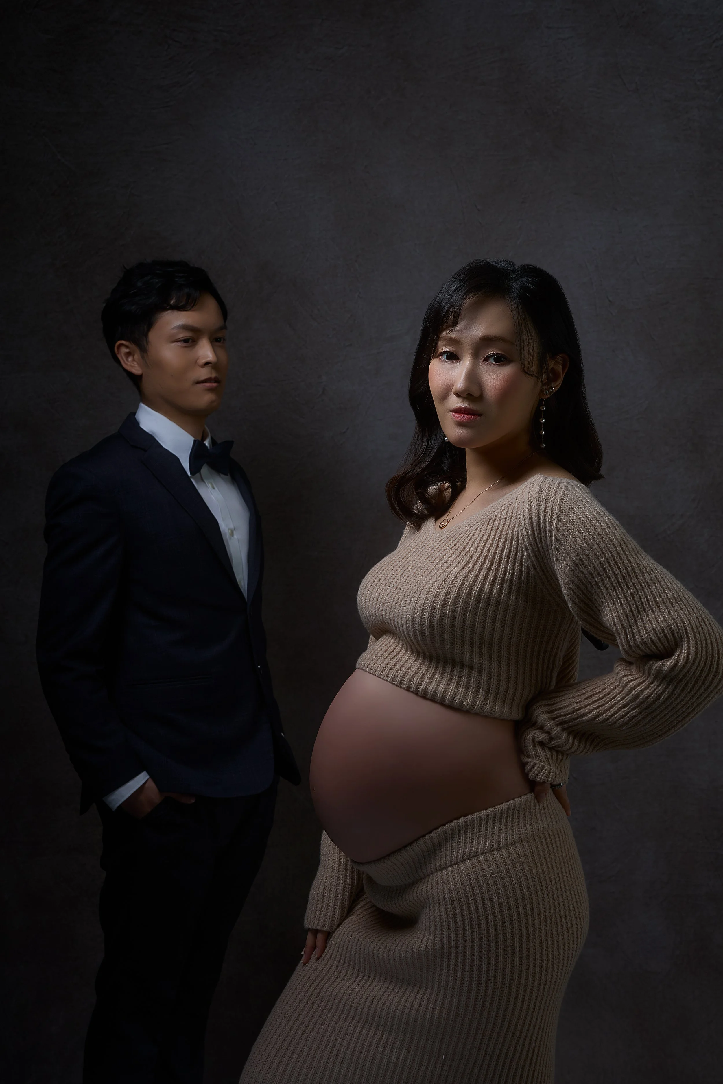 A pregnant woman poses with a serious expression, wearing a beige knitted crop top and skirt, with her hand on her hip, while a man in a tuxedo stands behind her looking at her, against a dark textured background.