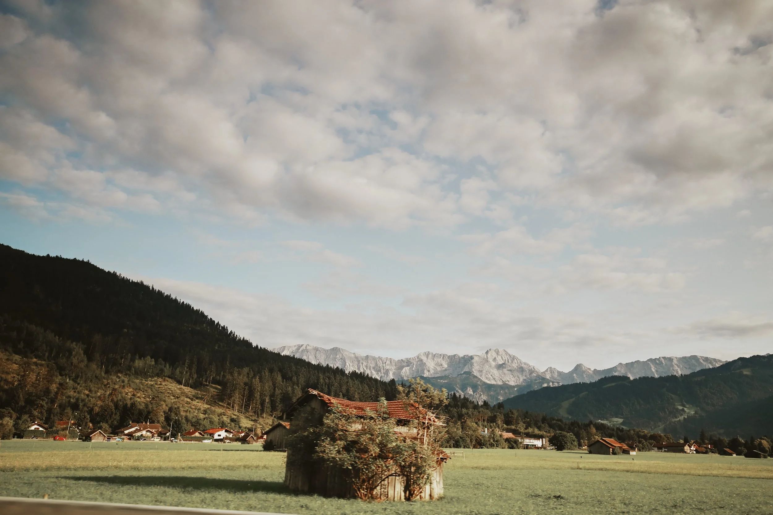 Scenic landscape with a green field, a small rustic barn, forested hills, and distant mountains under a partly cloudy sky.