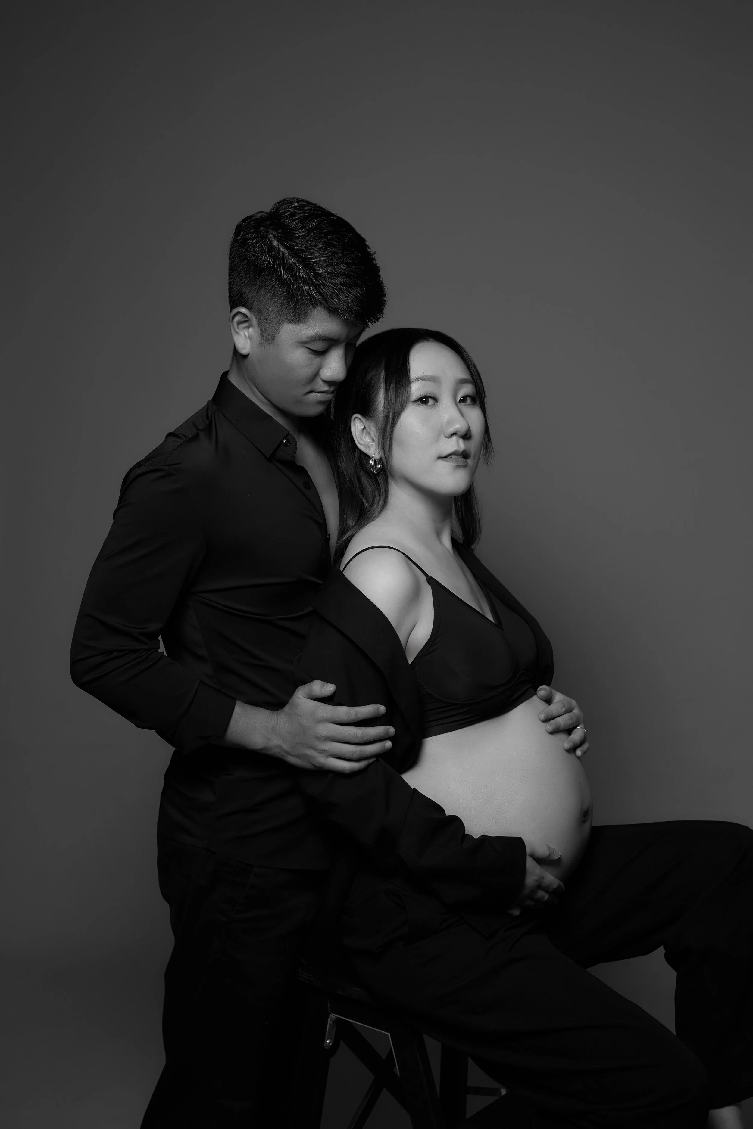 A black and white photograph of a pregnant woman sitting on a stool with a man standing behind her, both dressed in black clothing, with the woman wearing a bra and an open shirt. They are closely embracing, with the man's hand on her waist and her h
