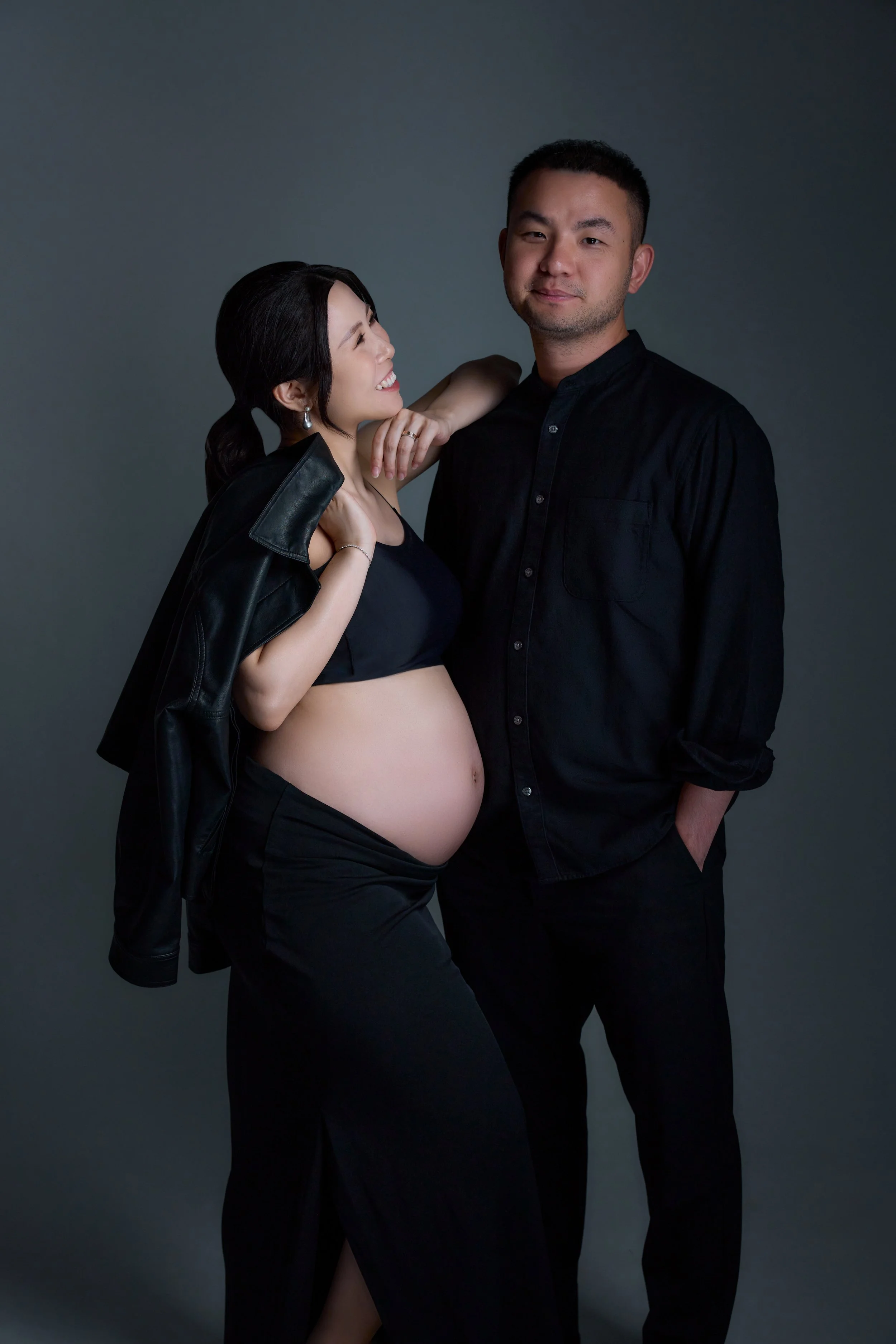 A pregnant woman and a man in black clothing stand close together, the woman smiling and looking at the man, with her hand on his shoulder, while the man looks straight at the camera with a serious expression.