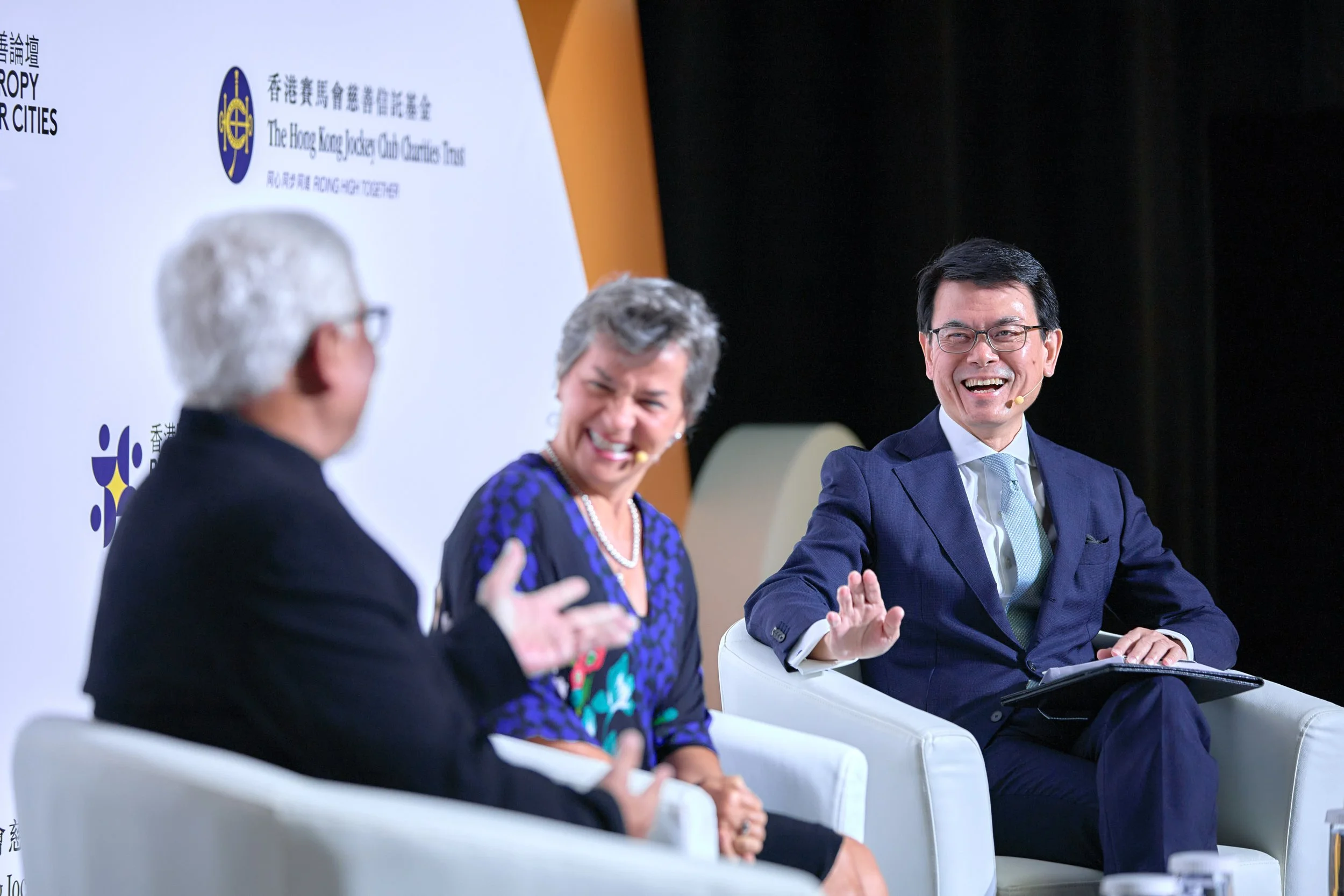 Three people in a panel discussion, smiling and laughing, seated on white chairs, with a backdrop featuring logos and text in both English and Chinese.