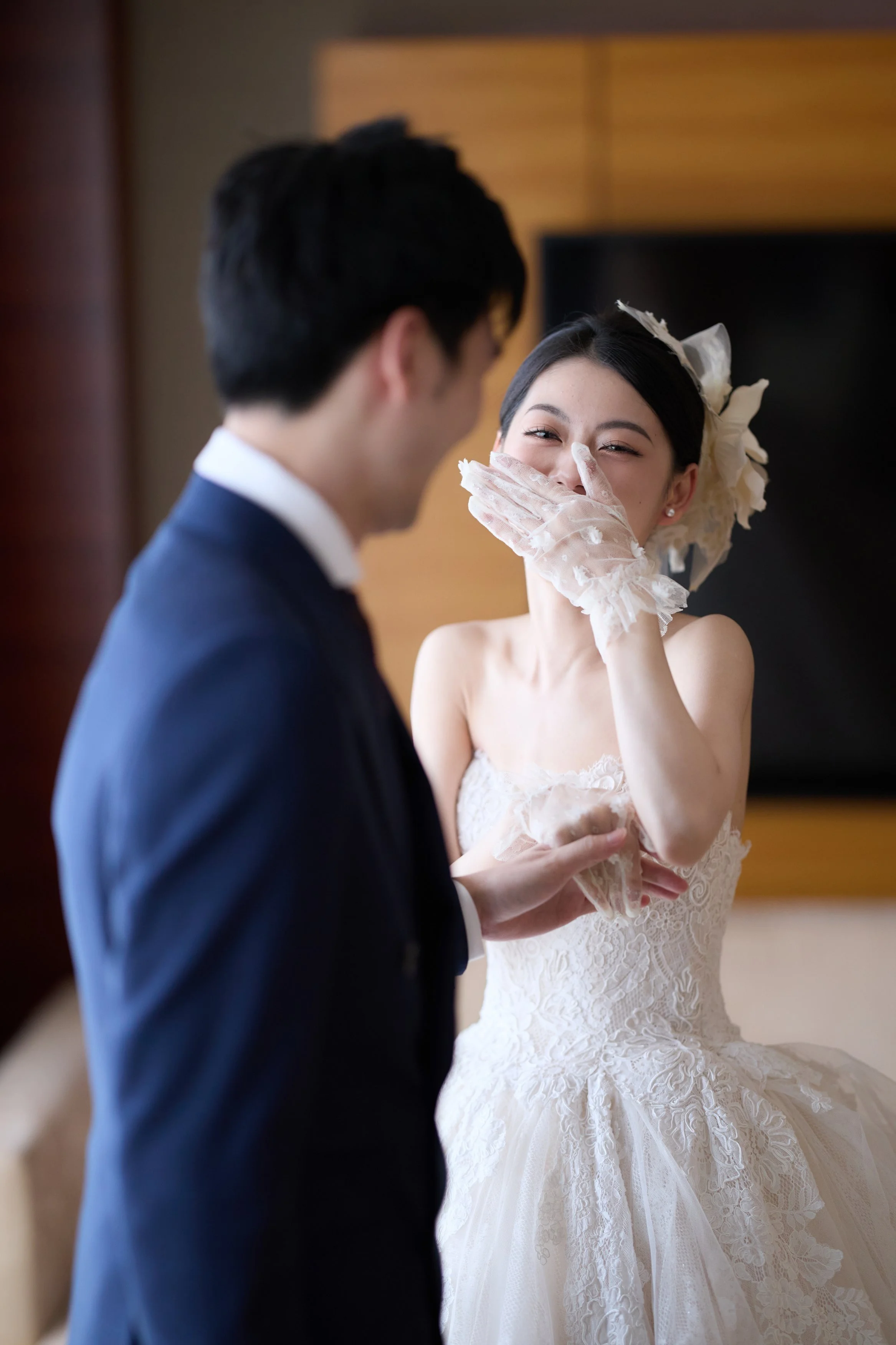 A bride laughing while covering her mouth with a lace glove, standing in front of a groom dressed in a navy suit, during a wedding celebration.