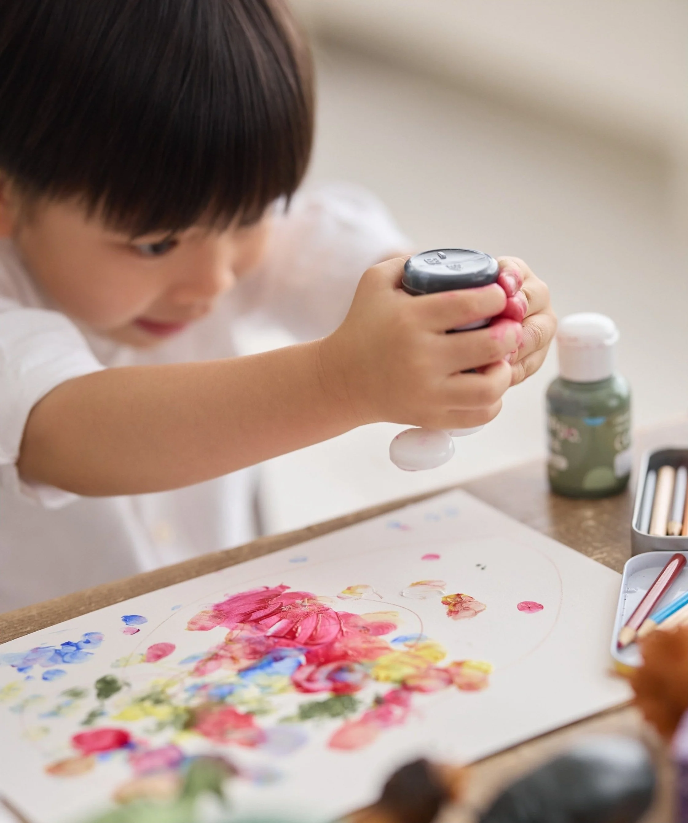 A young child with brown hair engaged in finger painting with pink, yellow, blue, and green paints on white paper, holding a black spray bottle.