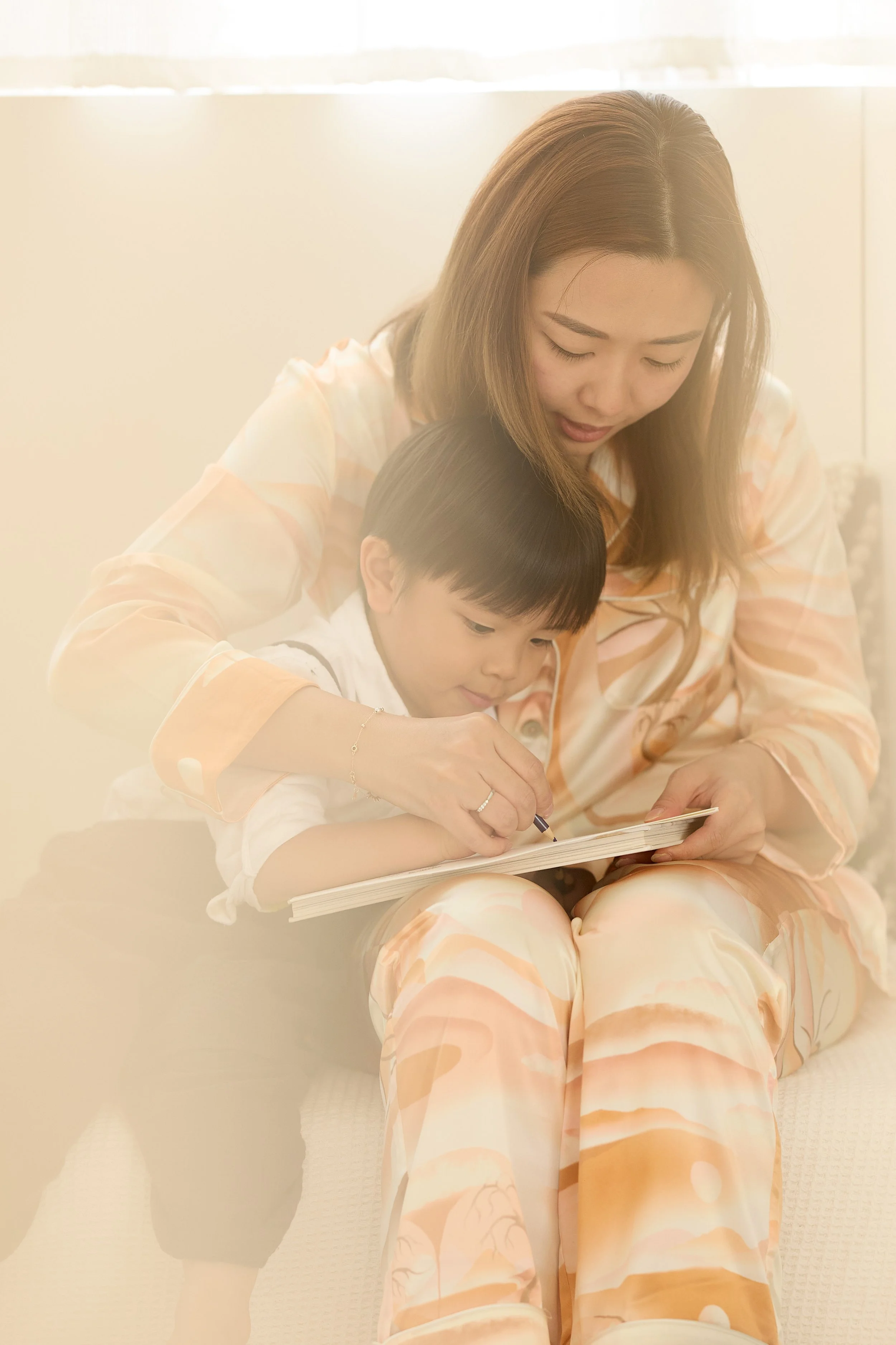 A woman and a young boy sitting together, reading a book. The woman is looking down at the book while the boy is focused on it, with the woman guiding his hand. They are indoors with soft sunlight coming through a window.