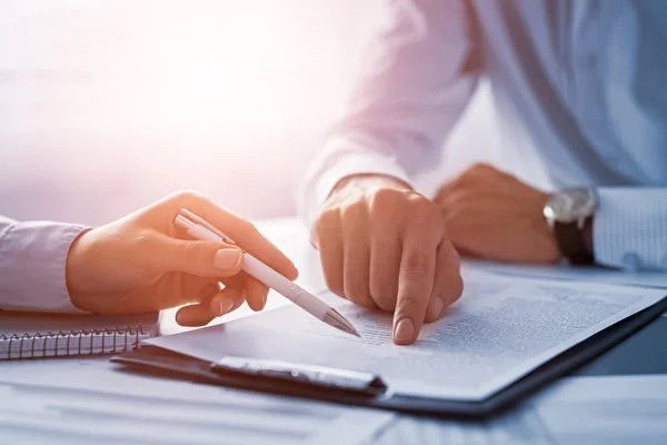 Two people sitting at a table reviewing documents, with one person pointing at a pen and document.