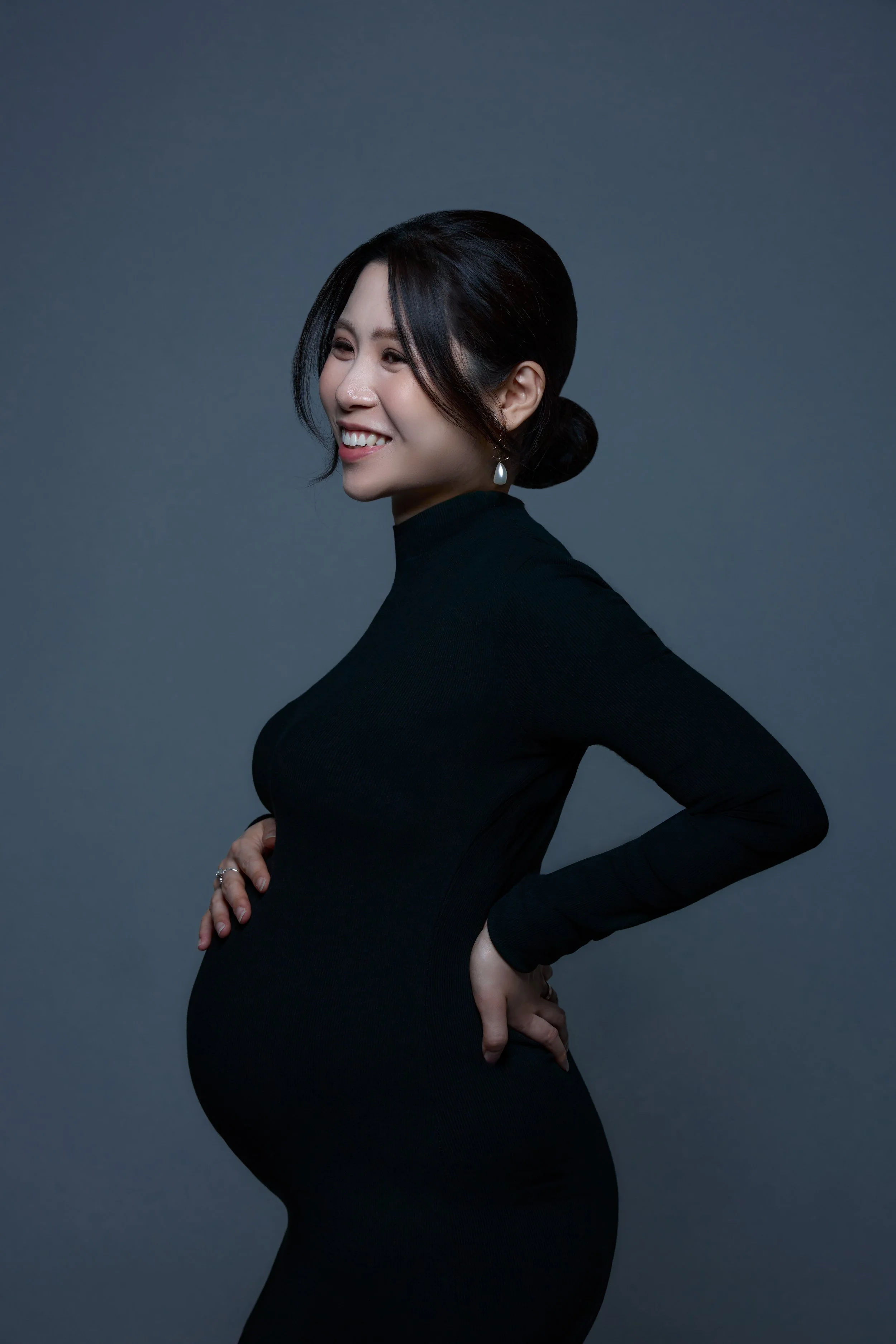 A pregnant woman in a black long-sleeve dress poses against a gray background, smiling and looking to the side.