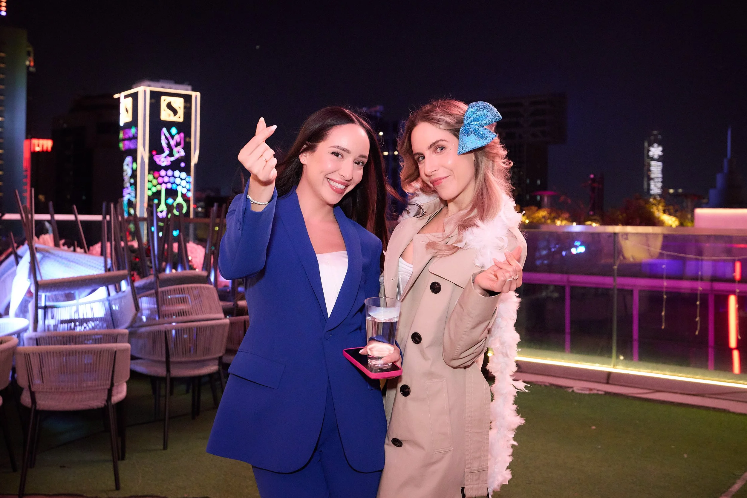 Two women smiling and posing together on a rooftop at night, with colorful city lights in the background. One woman is wearing a blue blazer and holding a glass of water, making a finger heart gesture. The other woman is wearing a beige coat with a feathered accessory and a blue bow in her hair, making a hand gesture and holding a pink phone.