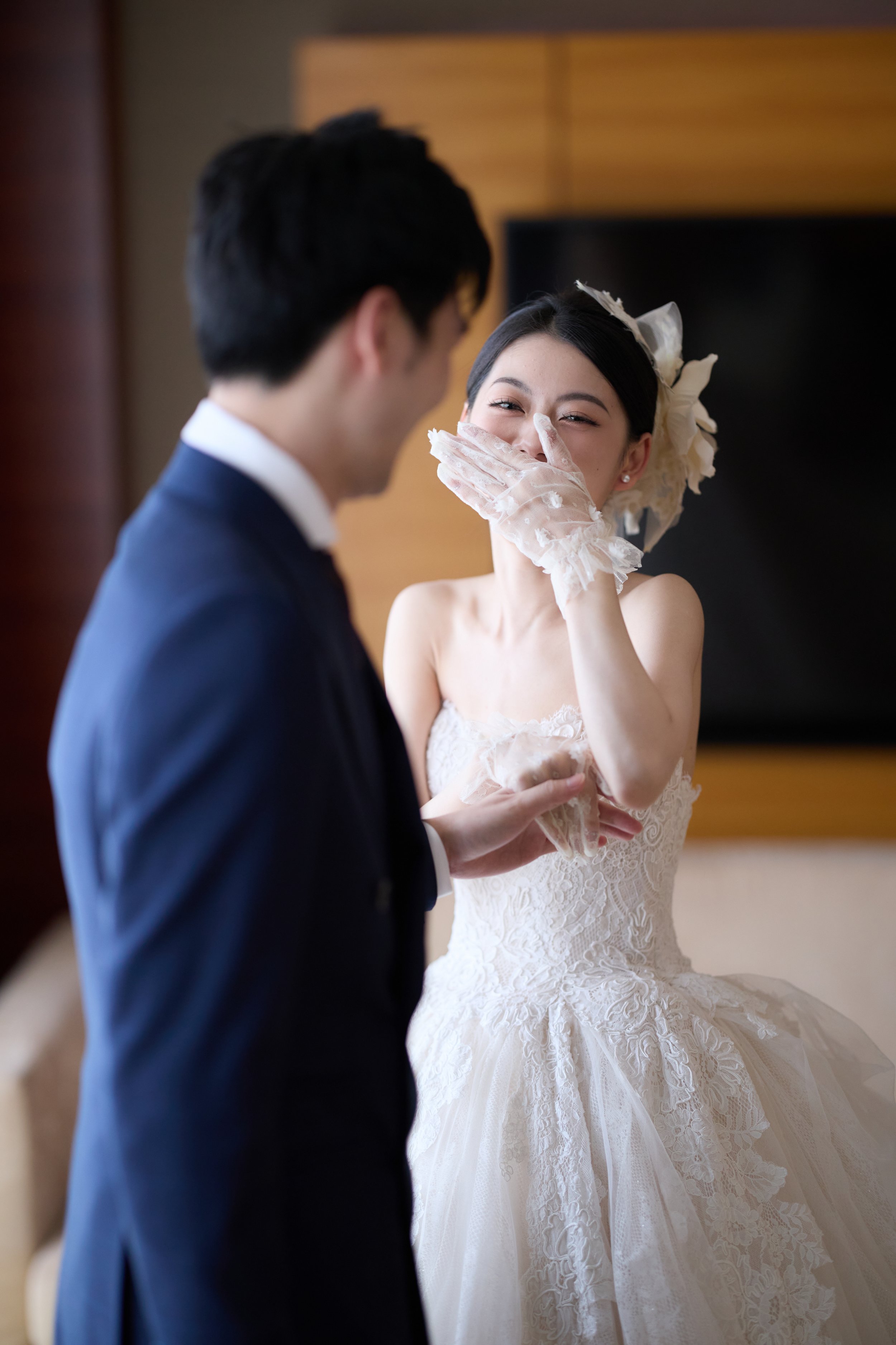 A bride in a wedding gown covering her mouth with her gloved hand while laughing at a wedding celebration.