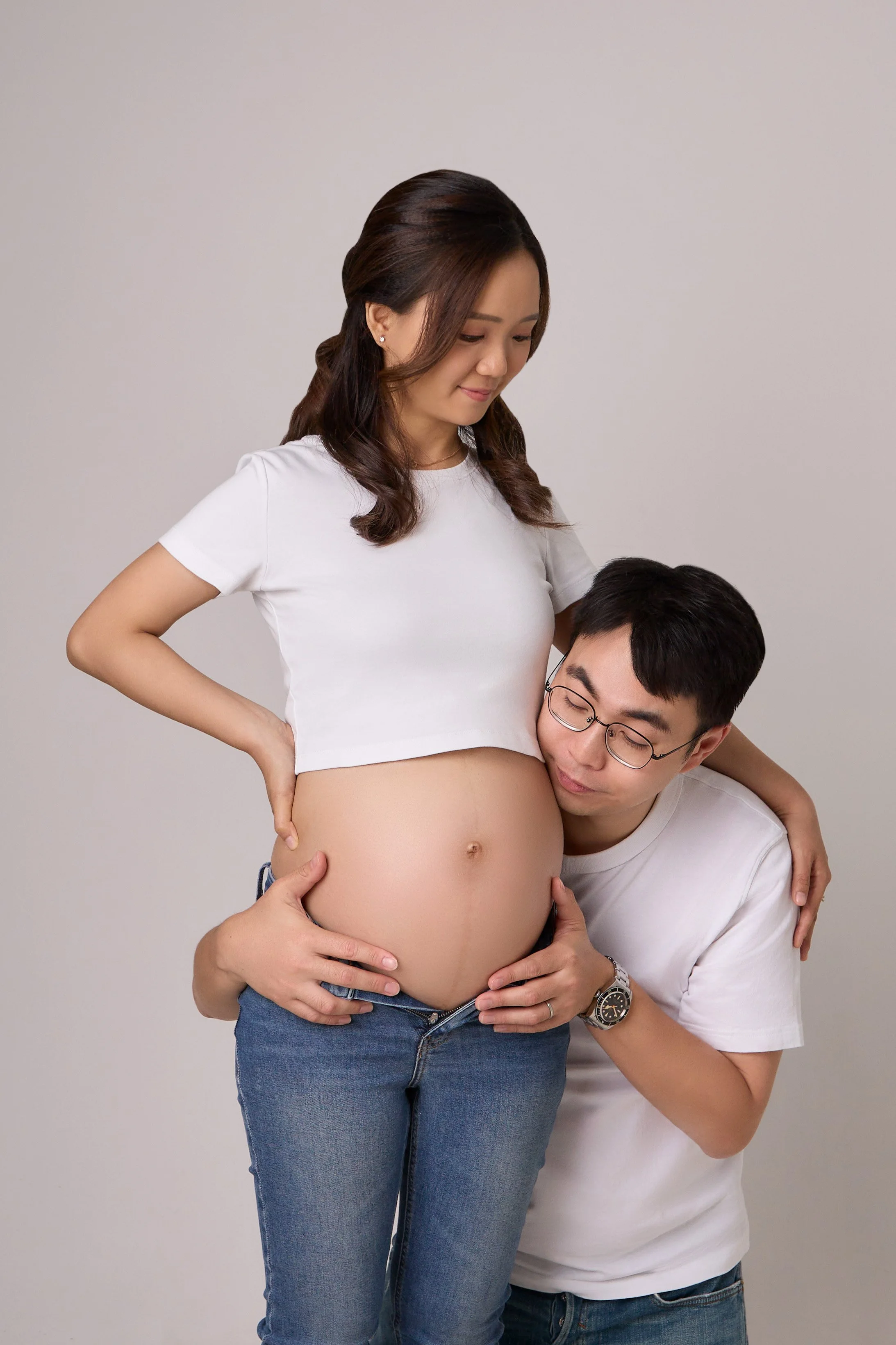 A pregnant woman in a white crop top and jeans is gently holding her belly, while a man in glasses and a white t-shirt is kneeling beside her, resting his head on her belly and holding it with both hands.