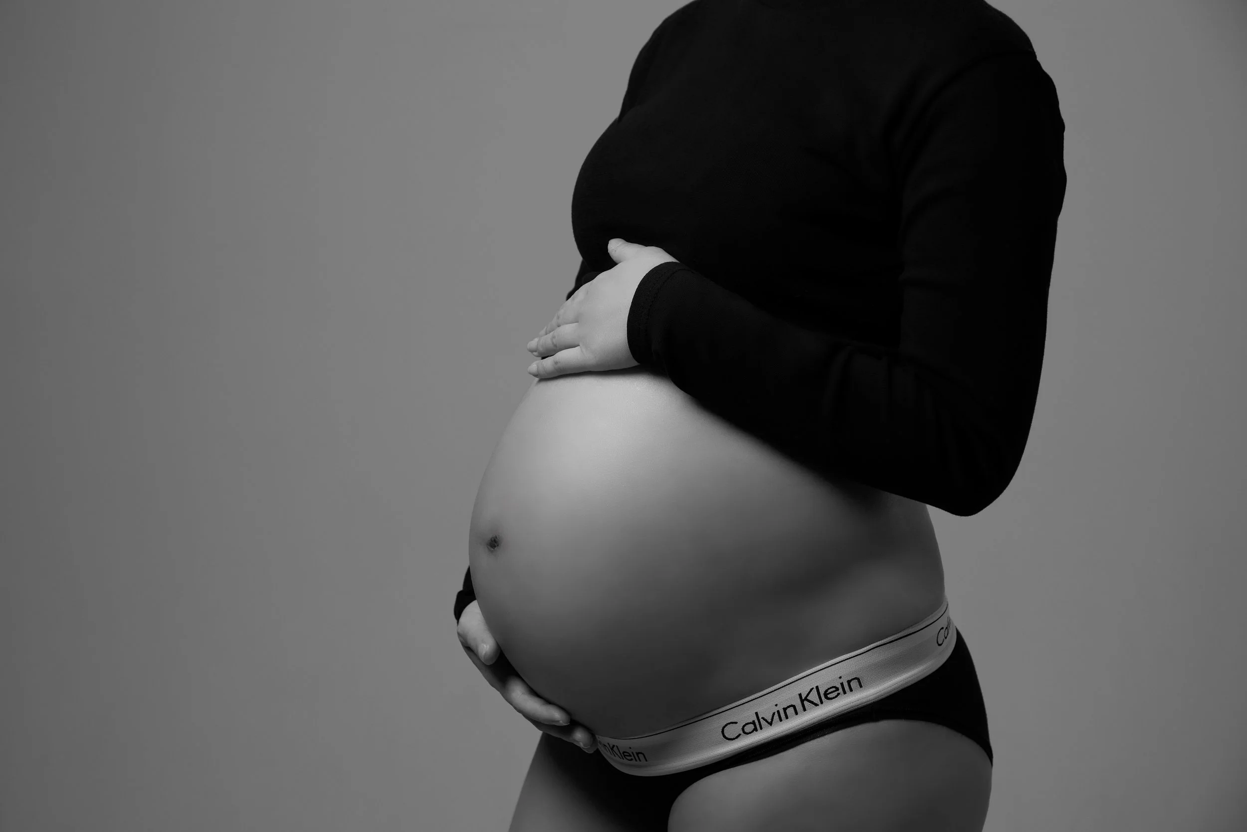 Black and white photo of a pregnant woman holding her belly, wearing black Calvin Klein underwear and a long-sleeve top.