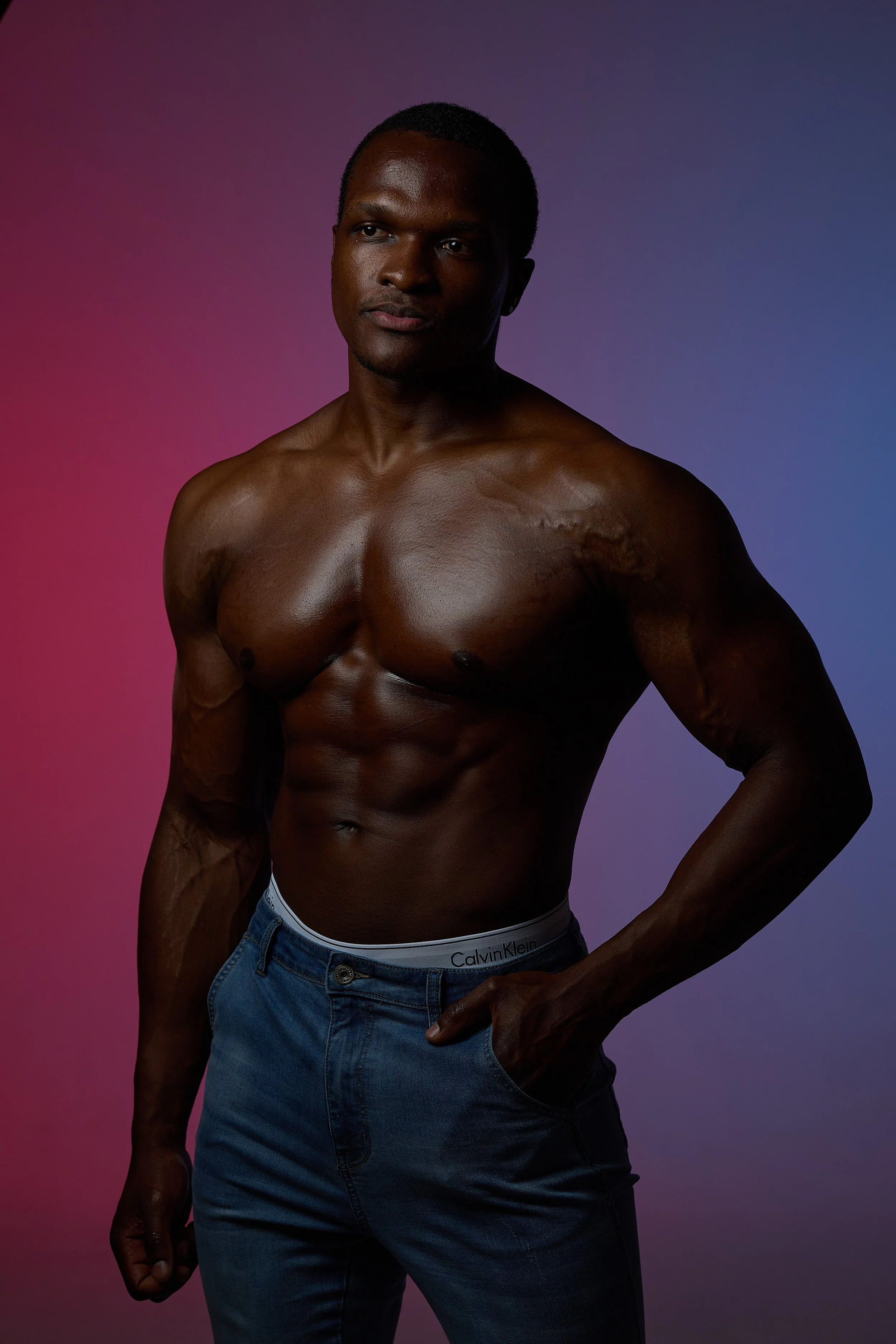 A fit, shirtless man with dark skin posing against a gradient background that transitions from pink to purple, wearing blue jeans and Calvin Klein underwear.