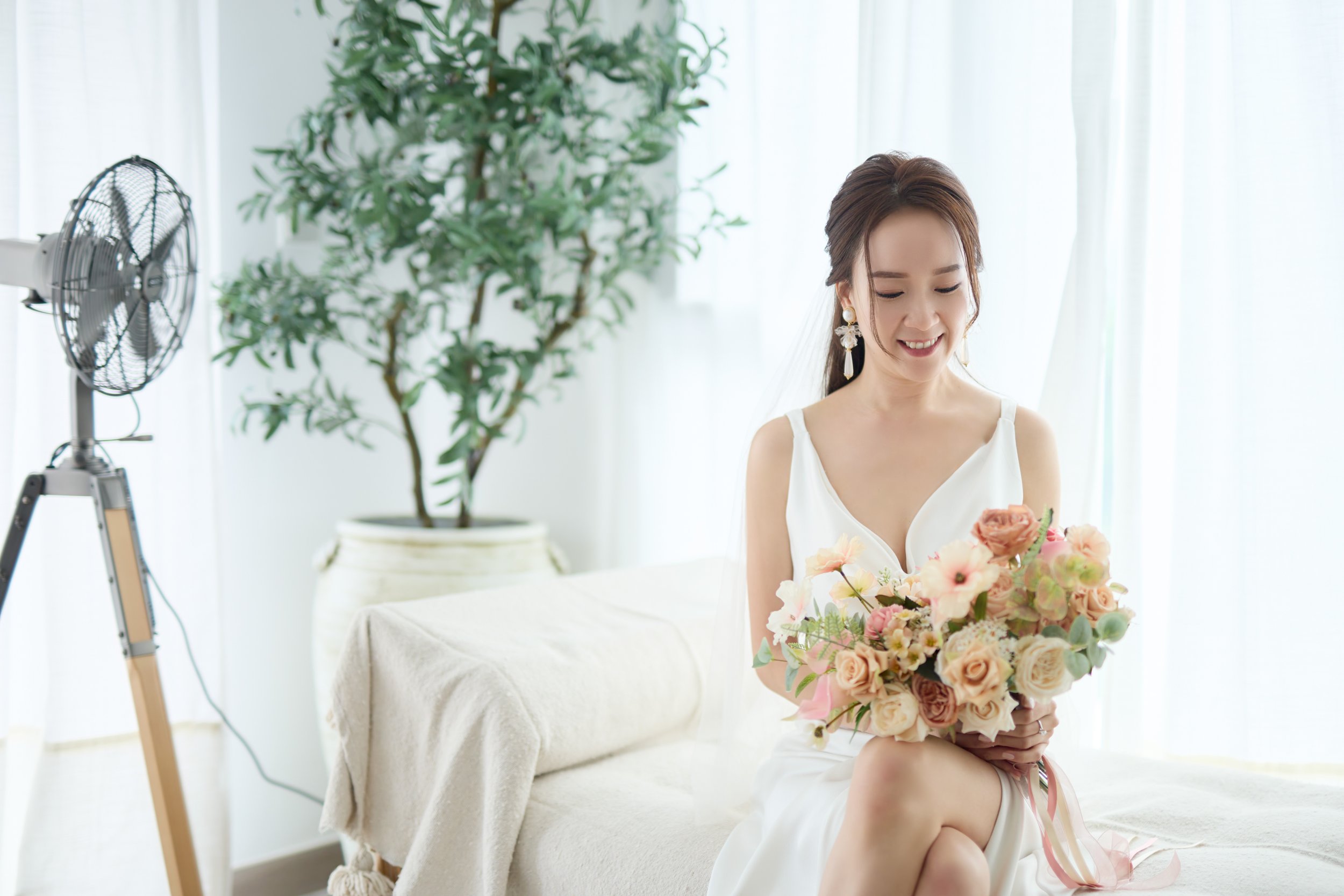 A woman in a white dress sitting on a sofa, holding a bouquet of pink and cream flowers, smiling while looking at the flowers.