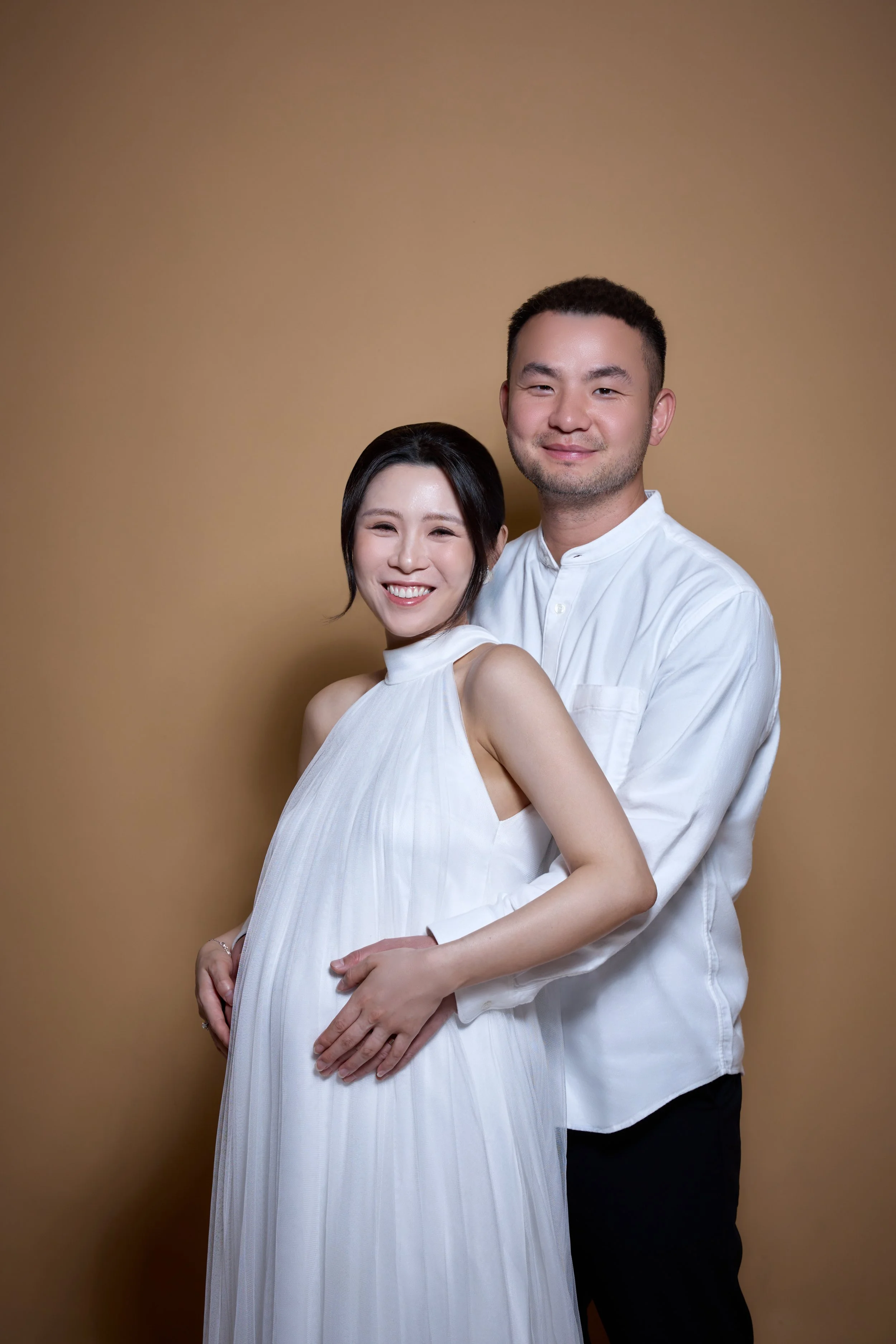 A smiling woman in a white dress and a man in a white shirt standing close together, with the woman holding her pregnant belly, against a plain brown background.