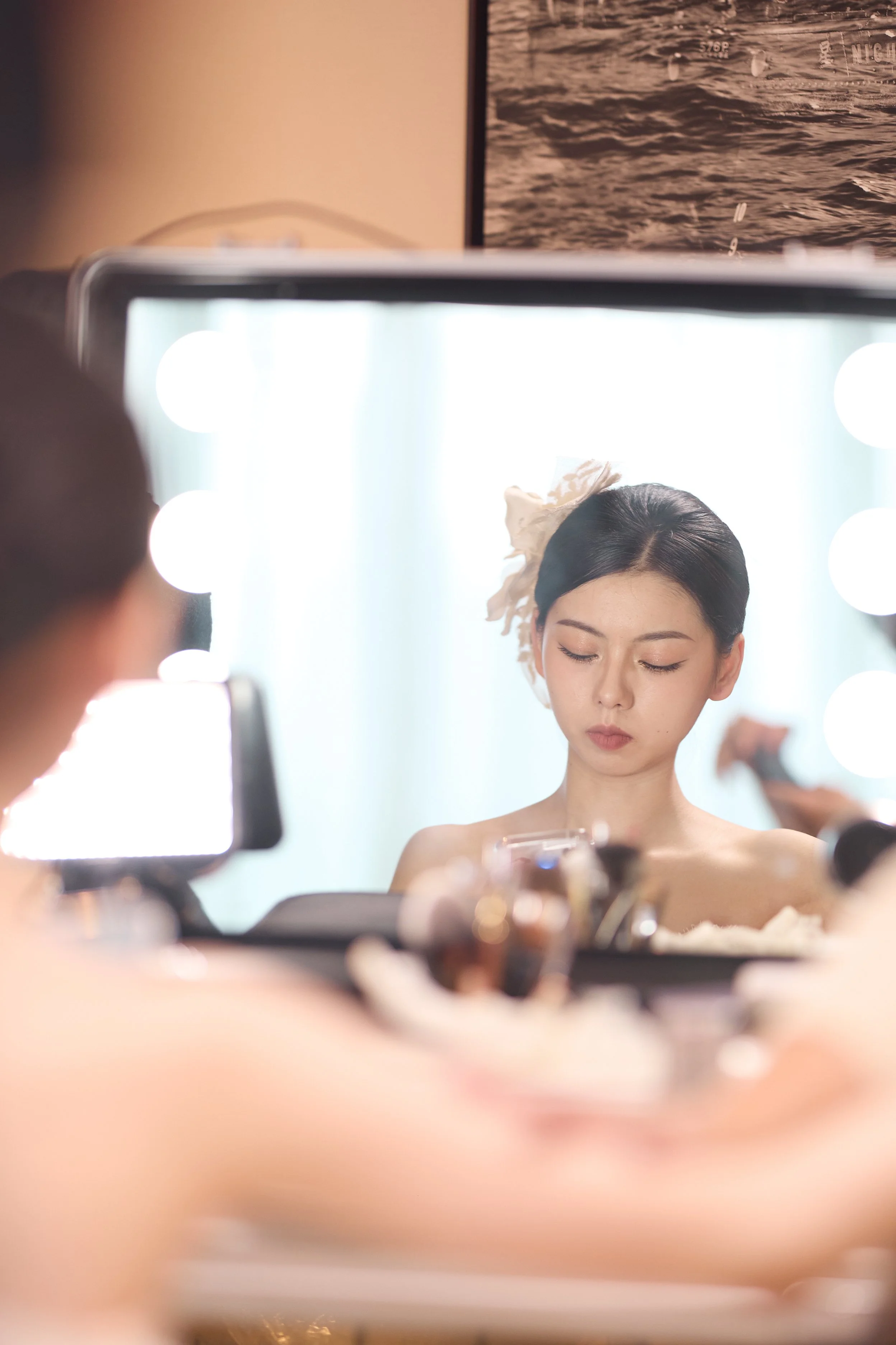 A woman getting her makeup done, seen through a mirror with bright lights, with her eyes closed and wearing a white floral headpiece.