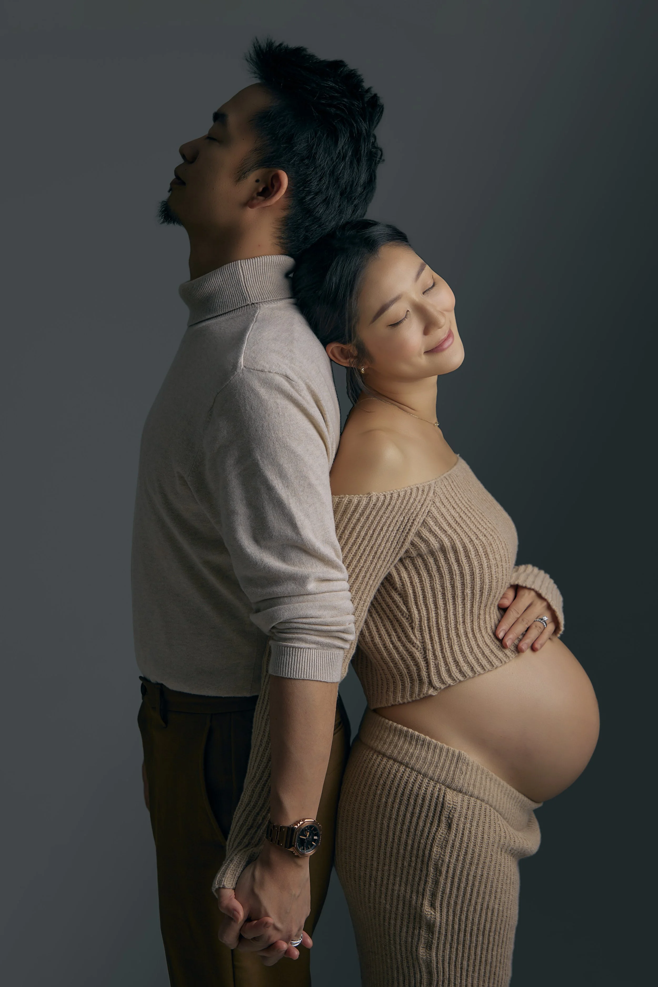 A pregnant woman and a man holding hands, both with eyes closed and smiling gently, standing back-to-back against a gray background.