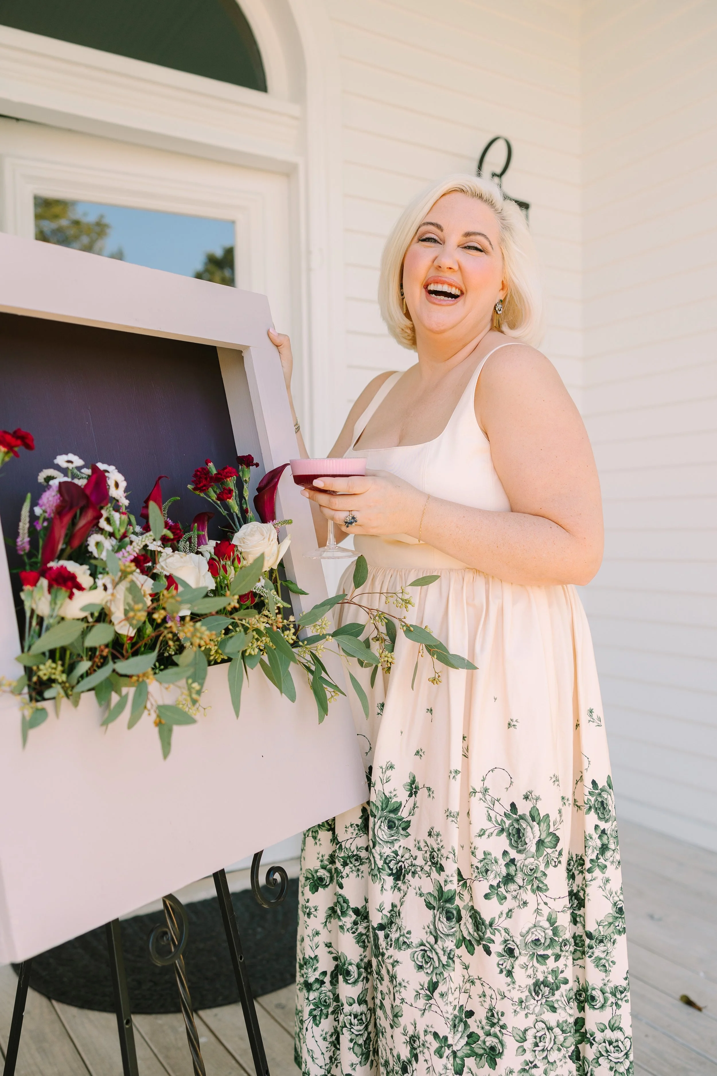A woman wearing a floral dress is smiling and holding a drink, standing next to a framed floral arrangement outside a white building.