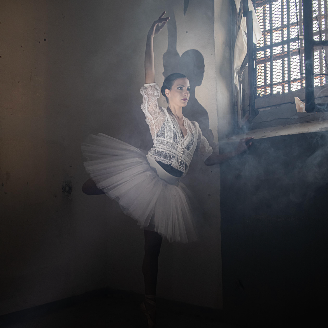Une ballerine en tutu blanc et blouse en dentelle pose dans une pièce sombre avec lumière venant d'une fenêtre.